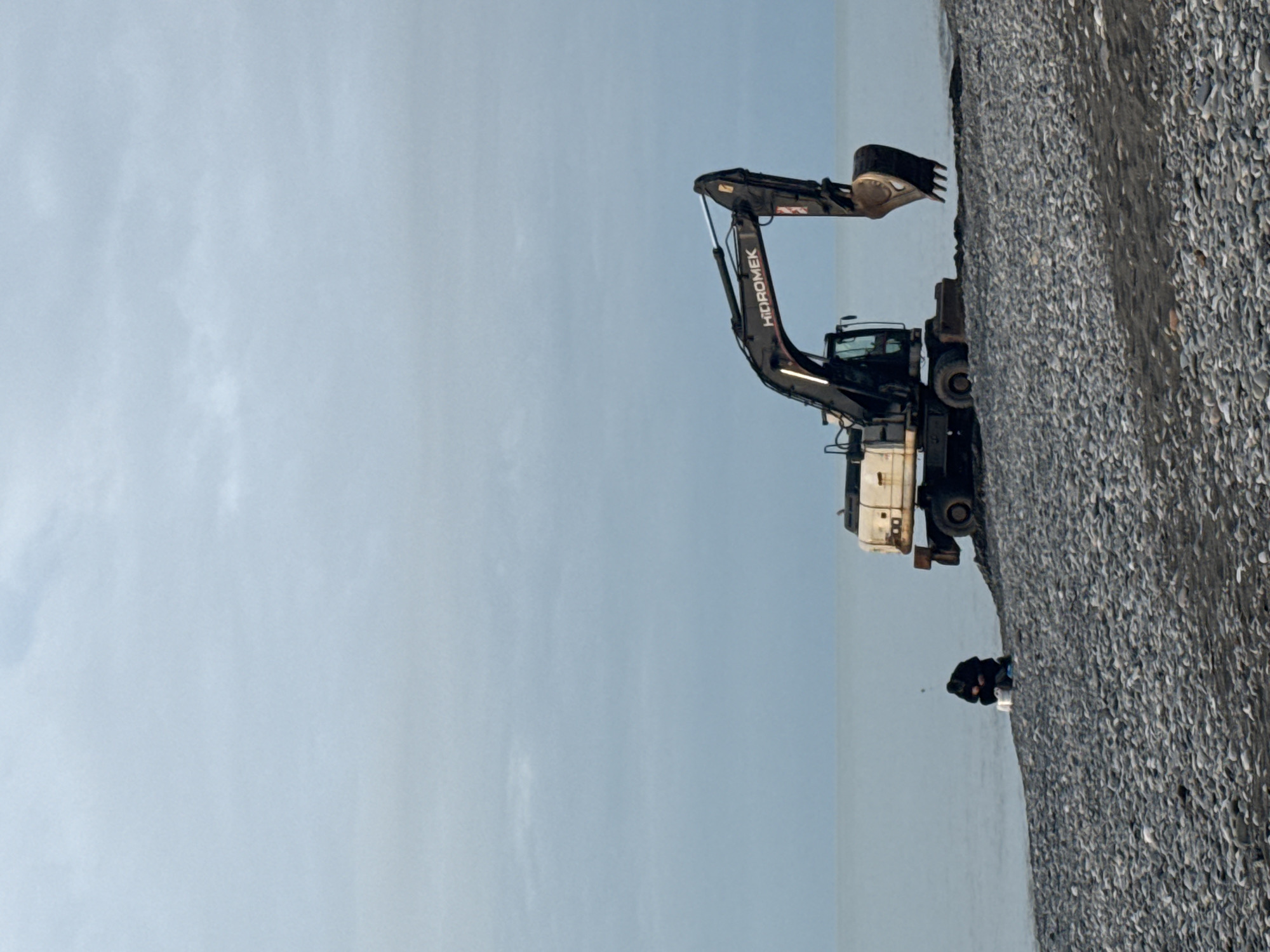 The image depicts a quarry in Batumi, Georgia. An excavator is positioned on the edge of a pile of gray aggregate material. A figure is observed near the base of the aggregate pile. The sky is a light, pale blue with few clouds.