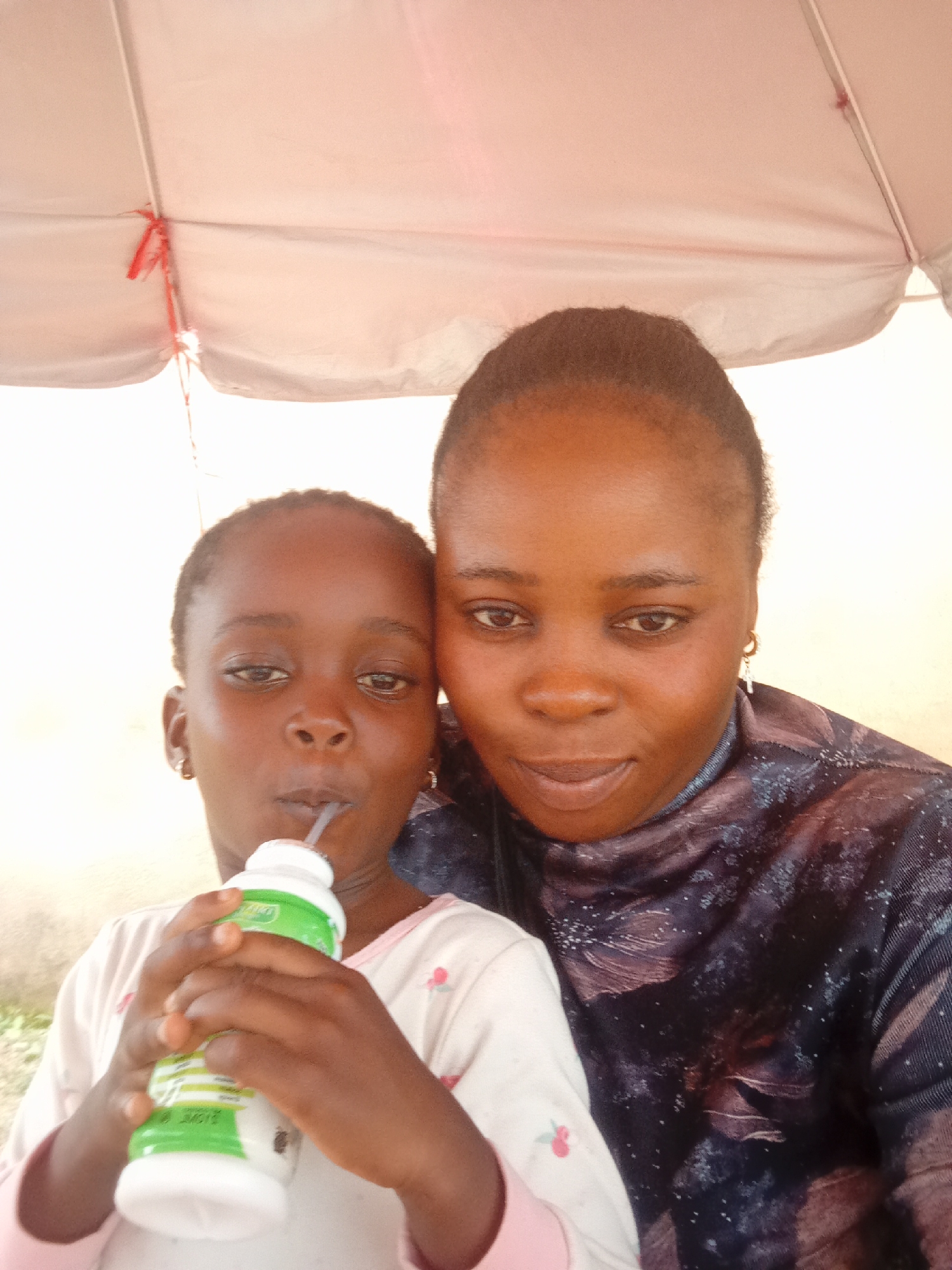 In Jos, Nigeria, a woman and a child are captured in a selfie taken outdoors, under the shade of a light-colored umbrella. The woman, on the right, has her head tilted slightly, and is looking towards the camera. She has dark skin, dark eyes, and is wearing a dark blue top with a patterned design. A gold earring with a cross pendant hangs from her right ear. To her left, the child, who is younger, is also looking at the camera and drinking from a white and green bottle with a straw. The child has dark skin, dark eyes, and is wearing a light pink shirt with a floral pattern. The background appears to be an outdoor setting with a neutral-colored wall. The lighting is bright, suggesting it is daytime.