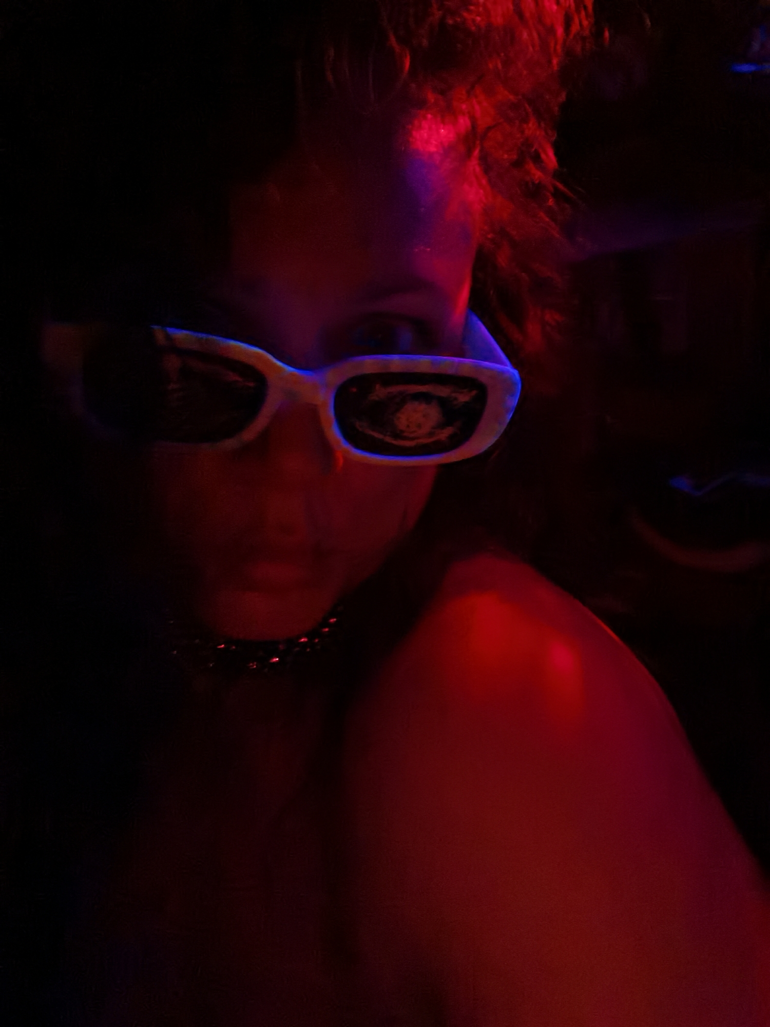 The image is a close-up, low-light photograph of a person wearing white-rimmed sunglasses and a choker necklace. The face is partially obscured by the sunglasses, which reflect an unknown image in the lenses. The lighting is primarily red and purple, casting strong shadows and highlighting parts of the person's face and shoulder. The hair appears dark and somewhat messy, with red highlights. The background is dark and indistinct, suggesting an indoor environment. There is no clear indication of the location being Chaikovsky, Russia.