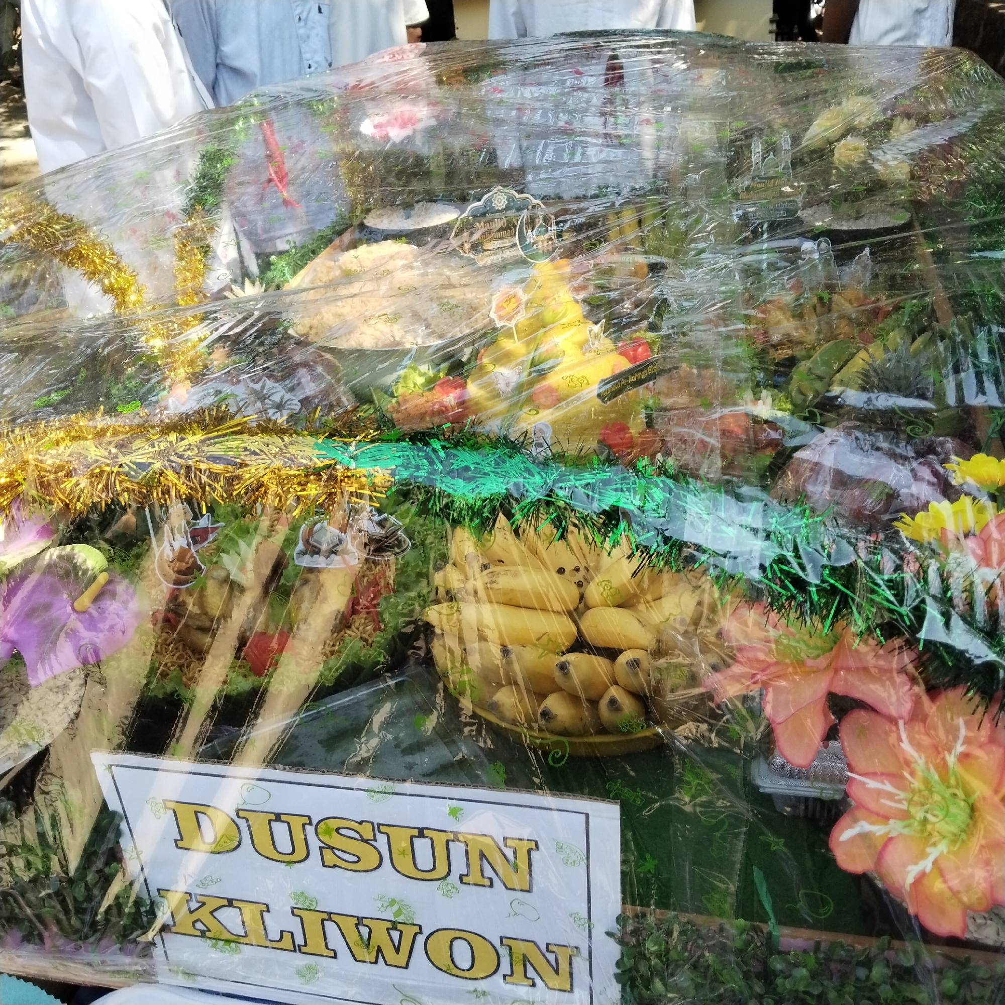 In Pamulihan, Indonesia, a close-up shot focuses on a rectangular food presentation, likely a tumpeng or similar festive dish, wrapped in clear plastic. The food is arranged in layers and adorned with decorative elements like golden and green tinsel, artificial flowers in shades of pink and orange, and what appear to be small decorative figurines. Visible food items include bunches of bananas, leafy green vegetables, and other unidentifiable food items presented in conical shapes. A white sign with bold yellow lettering is prominently displayed at the bottom, reading "DUSUN KLIWON." The background shows parts of people's bodies, suggesting the presentation is part of a gathering or event. The lighting is bright, creating reflections on the plastic wrap.