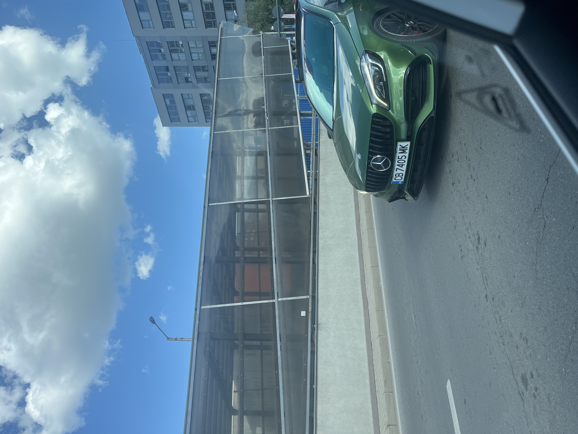A green Mercedes-Benz A-Class is driving on a city street in Sofia, Bulgaria. The car has a license plate that reads "CB 7405 MK". In the background, there is a modern building with many windows and a covered walkway with transparent panels. The sky is blue with some white clouds. The street is paved and has a white lane marking. The image is taken from inside another vehicle, as indicated by the reflection in the foreground.