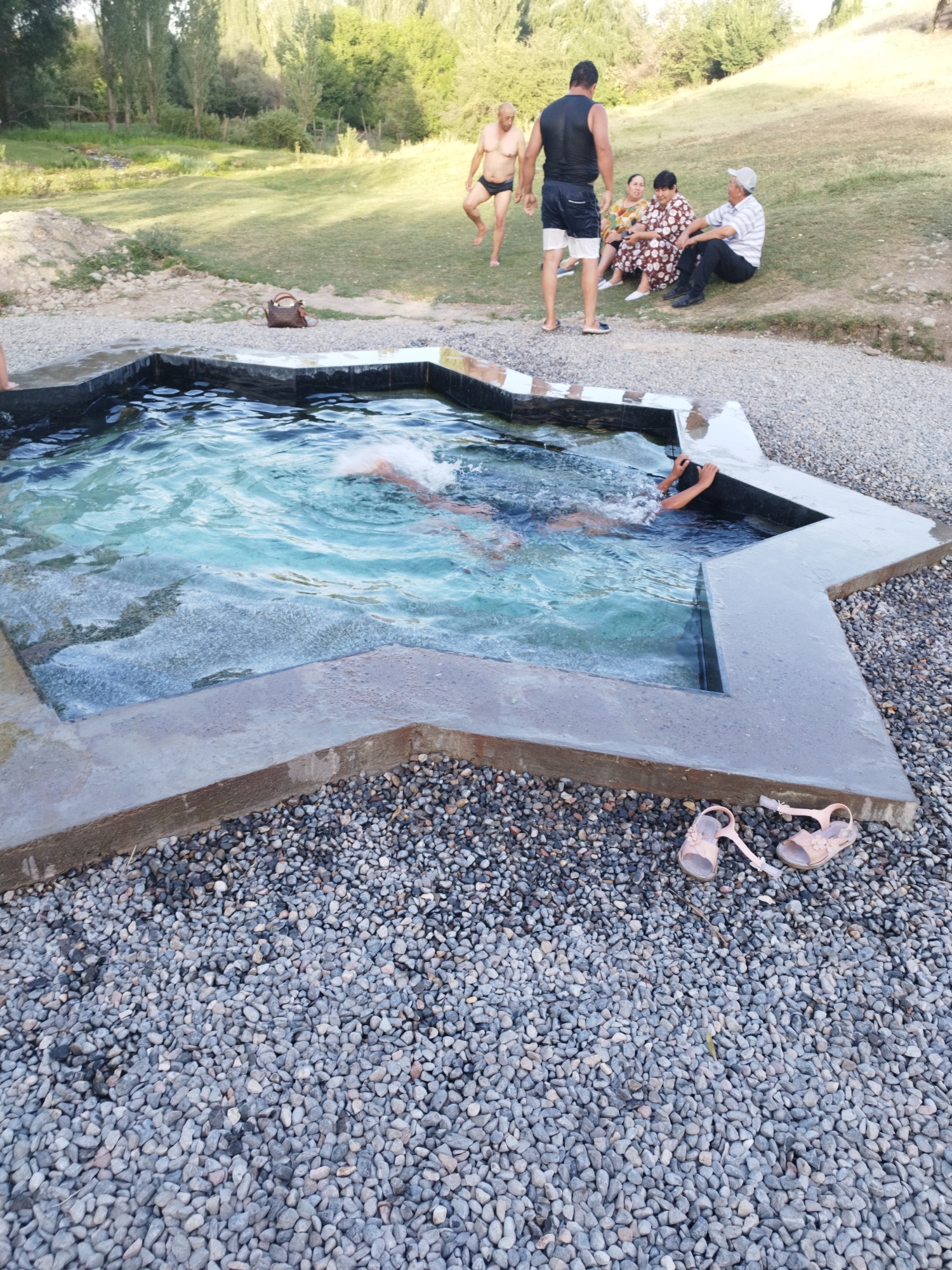 In Көксаяқ ауылдық округі, Kazakhstan, a group of people are gathered around a unique, star-shaped pool of water. The pool has a concrete border with a dark tile inset. Three individuals are seated on a grassy incline to the right of the pool: a woman in a patterned dress and a man in a striped shirt and cap, who appear to be conversing. Another woman in a floral dress is seated next to them. To the left, a man in swim trunks is standing, and another man in black shorts and a black tank top is standing by the edge of the pool. Two individuals, appearing to be children, are partially submerged in the water, with one clearly visible being supported by their hands on the pool's edge. A brown handbag rests on the gravel path near the pool. In the foreground, two light-pink children's sandals lie on the gravel. The background consists of a grassy field with scattered trees and foliage, suggesting an outdoor recreational area.