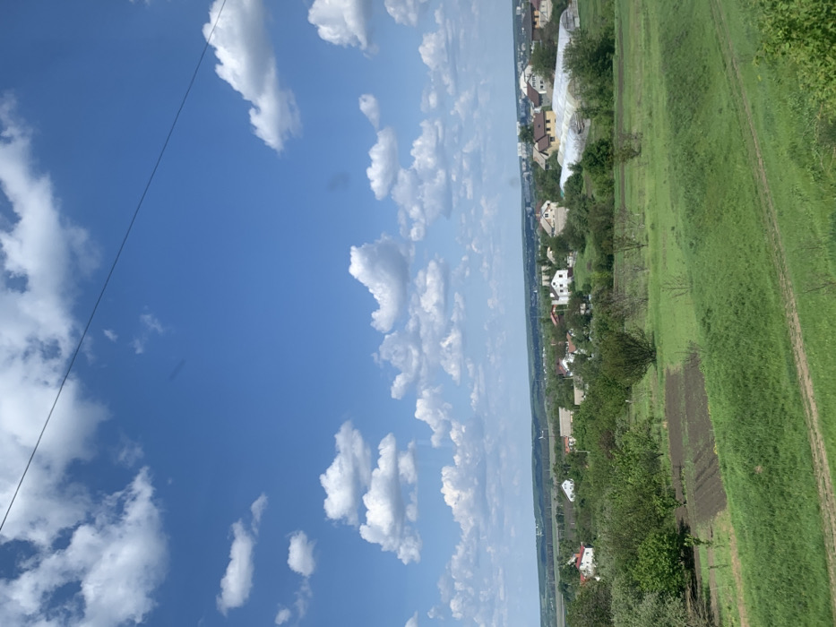 The image shows a panoramic view of a suburban area in Korotych, Ukraine, located at latitude 49.956 and longitude 36.031. The scene is dominated by a bright blue sky with scattered white cumulus clouds. In the foreground, a rolling green field with a dirt path stretches towards a cluster of trees and houses. The houses are mostly single-story structures with visible white walls and red roofs. A few greenhouses can be seen in the distance, suggesting agricultural activity. There are no people present in the scene. The overall atmosphere is peaceful and idyllic.