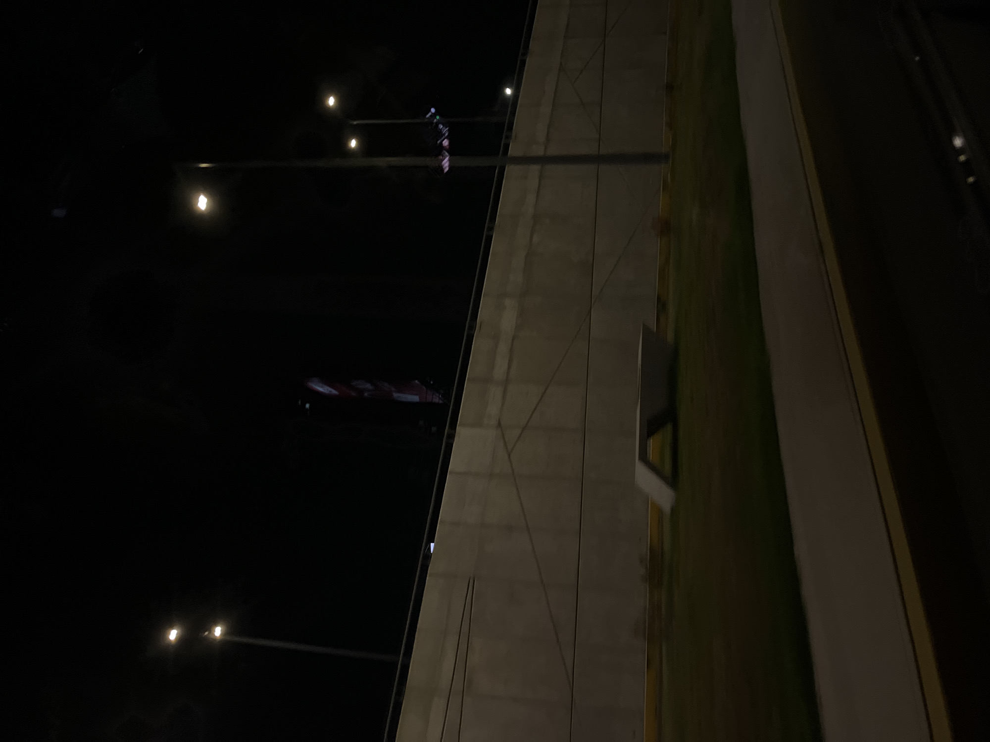 The image shows a night scene in Puebla, Mexico. A tall, concrete building with geometric patterns stands next to a green area with a white stripe. Streetlights illuminate the surroundings, and a distant building with red and white lights can be seen in the background.