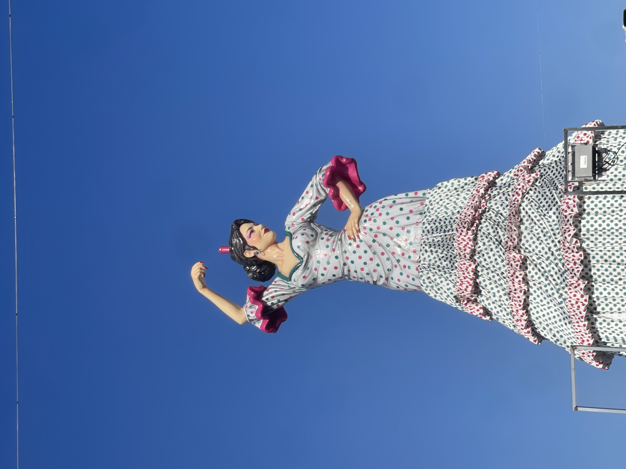 The image shows a large figure of a woman in a traditional flamenco dress against a clear blue sky. The dress is predominantly white with red and blue polka dots, adorned with ruffles. The woman's hair is styled in a bun with a red flower accessory. Her pose suggests she is dancing, with one arm raised. There is a metal support structure and a power box visible near the bottom right corner of the frame. The scene is likely captured outdoors, possibly during a festival or event in Barcelona, Spain, where such figures are common decorations.