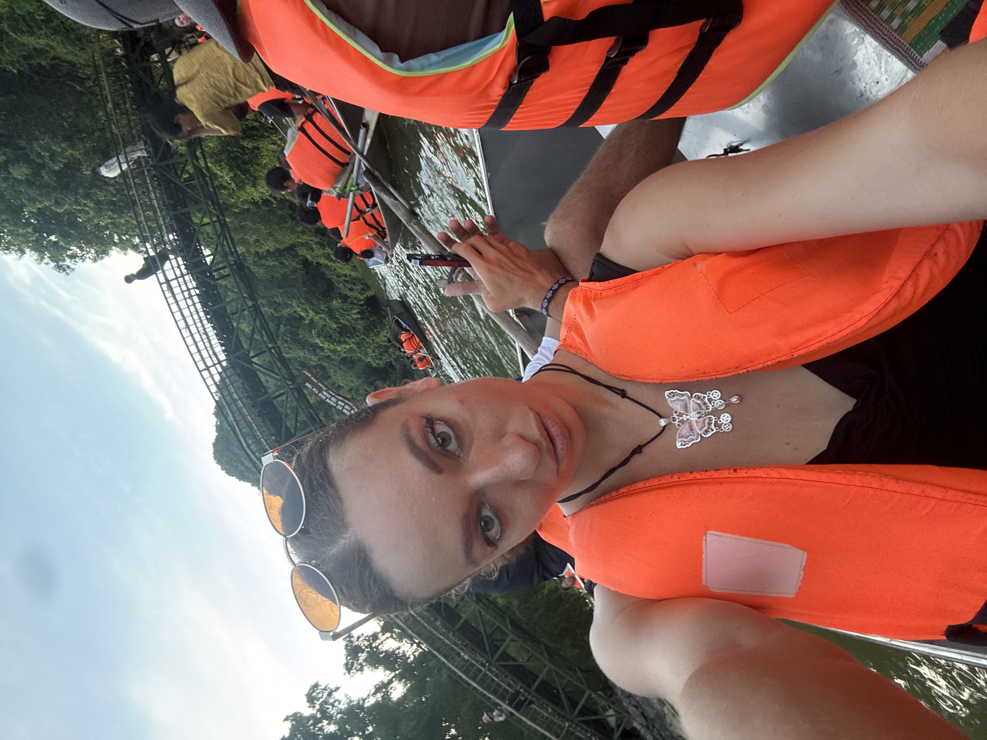 A woman is taking a selfie in a boat on the water, wearing a bright orange life vest and sunglasses. She has on a butterfly pendant necklace. In the background, other people in life vests are also in boats, and a wooden structure resembling a bridge or walkway is visible over a lush green, treed hillside. The scene appears to be a tourist activity on a river or lake in a natural setting. The presence of boats, life vests, and the mountainous terrain suggests this location could be near the karst landscape of Ninh Bình province, possibly within Yên Sơn Ward, Vietnam.
