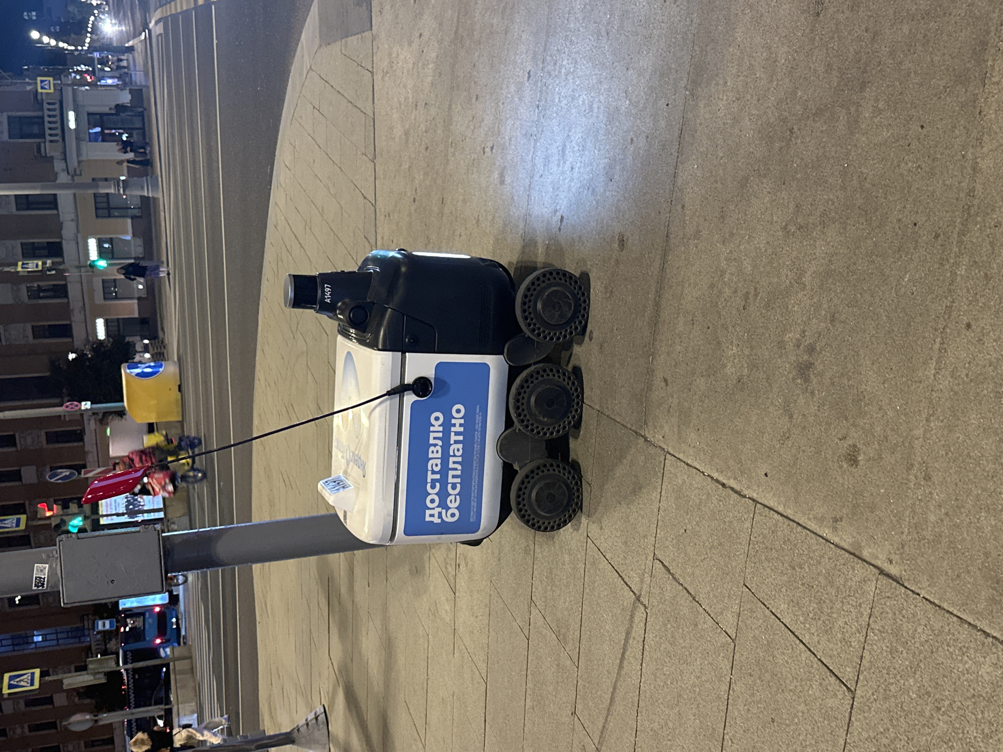 The image shows a Yandex.Lavka delivery robot on a sidewalk in Moscow, Russia. The robot is white and black, with a blue sign on the side that reads "доставлю бесплатно" (delivery free). It has four wheels and a black antenna. In the background, there is a street with cars and pedestrians, and buildings. The overall scene appears to be at night or dusk, as indicated by the artificial lighting.