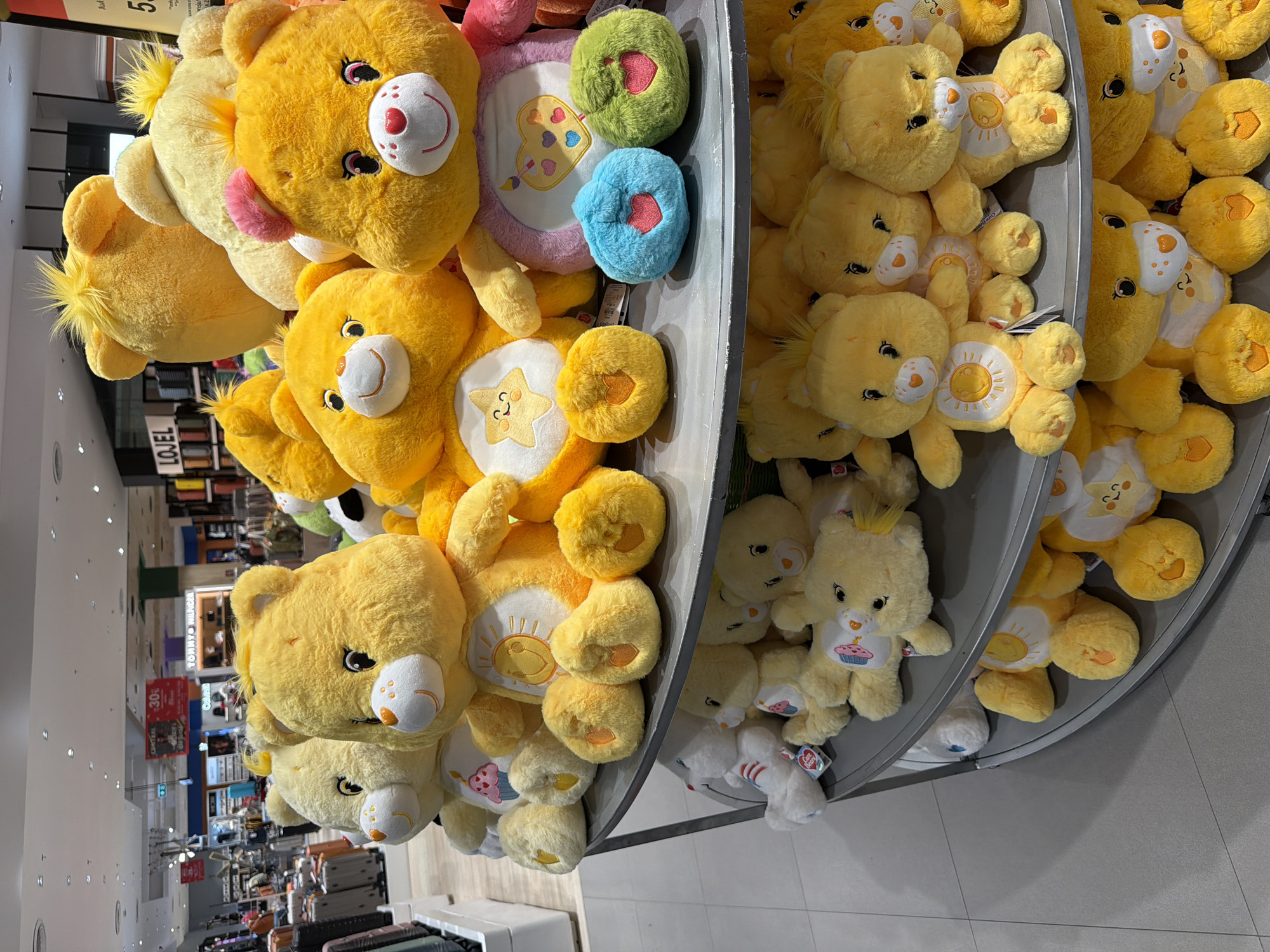 The image depicts a multi-tiered display stand in a store, likely in Baan Chaweng, Thailand, although no location clues are visible in the image itself. The shelves are filled with yellow plush Care Bears. The bears are various shades of yellow, with differing belly designs (sun, star, painter's palette). The store's interior is visible in the background, showing clothing racks, luggage, and signage.
