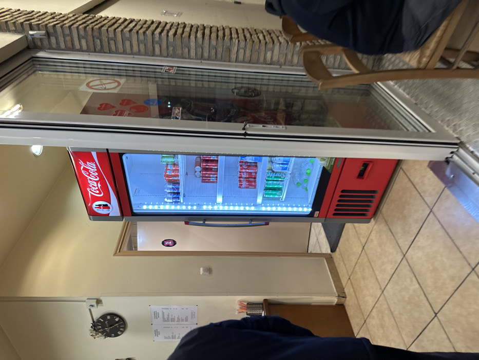 The image captures an indoor scene, likely inside an eatery or cafe in Nea Erithrea, Greece. The focal point is a red Coca-Cola branded display refrigerator, brightly illuminated from within, showcasing rows of soft drink cans. Visible brands include Coca-Cola (red cans), Fanta (orange cans), and Sprite (green cans), neatly arranged on multiple shelves. The refrigerator is seen through a wide glass entrance door or partition, which also reflects parts of the interior space.
Above the glass door, there is a section of exposed brickwork, suggesting a rustic architectural element. Inside the reflection on the glass, a circular "no smoking" sign is clearly visible, along with a blurred reflection of red shapes, possibly decorative hearts or logos, and the outline of a person seated at a table.
To the right of the glass door, a wooden chair is visible, indicating a seating area. The floor is covered with light-colored square tiles. To the left, the interior wall is painted a light yellow or cream color. Further inside the establishment, a wall-mounted clock with Roman numerals and ornate hands shows the time to be approximately 10:15 or 10:20. Below the clock, several sheets of paper, likely menus or informational notices, are affixed to the wall.
In the bottom right corner of the frame, the shoulder and arm of a person wearing a dark blue jacket or shirt are partially visible, suggesting the photograph was taken from a seated position. The overall impression is that of a casual, well-lit commercial space during operational hours. No specific activities are ongoing in the captured moment, presenting a static snapshot of the interior.