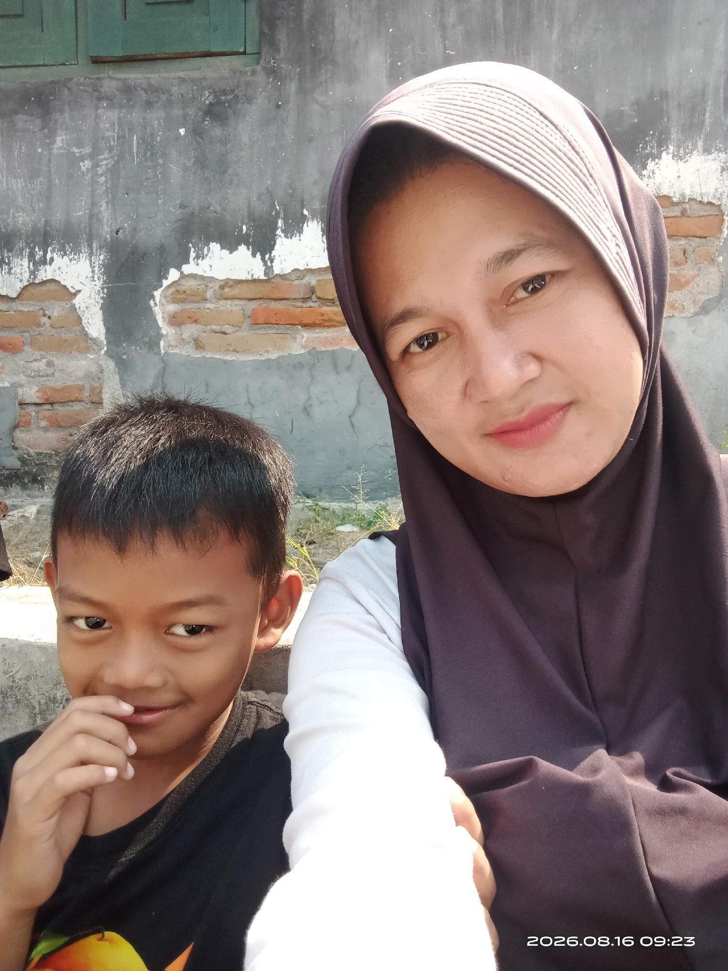 A medium shot captures a woman wearing a dark purple hijab and a white shirt posing for a selfie with a young boy in a black t-shirt. The boy is to the left of the woman, with his right hand near his mouth, and he is looking at the camera with a slight smile. The woman is on the right, also looking at the camera with a gentle smile. The background shows a distressed, weathered wall with exposed bricks and green window frames. The scene appears to be outdoors in a possibly rural or older urban setting in Banyuarang, Indonesia. The bottom right corner of the image displays a timestamp: 2026.08.16 09:23.