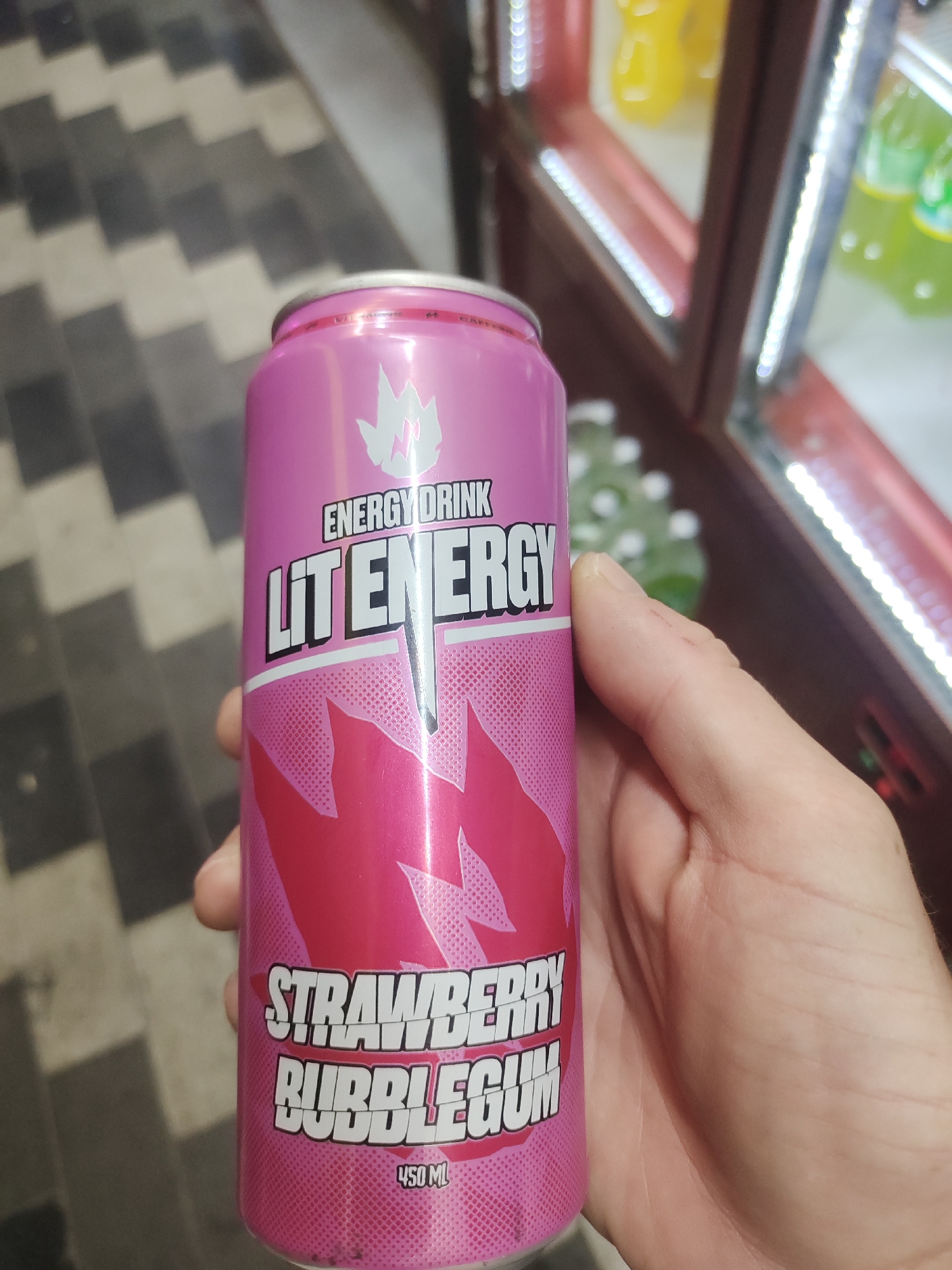 An image shows a person's hand holding a pink aluminum can of "LIT ENERGY" strawberry bubblegum flavored energy drink. The can has white text and a flame logo. The background appears to be a checkered floor and a vending machine with bottles of drinks inside. The image is taken in Tashkent, Uzbekistan.