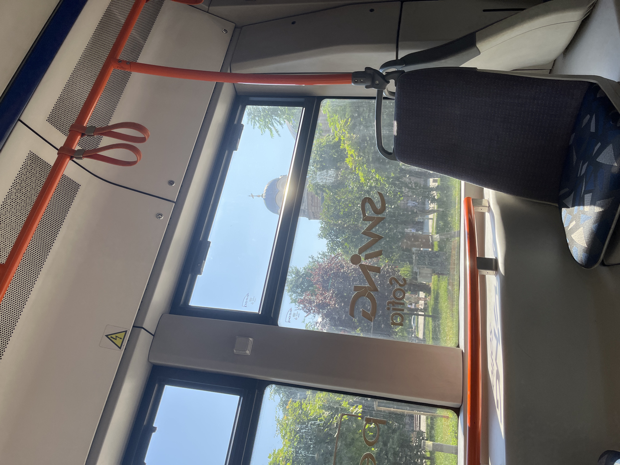 The image shows the interior of a public transport vehicle, possibly a tram or bus, in Sofia, Bulgaria. Through the window, a church with a dome and a cross is visible, surrounded by trees and a blue sky. On the window, the word "SWING" is reflected backwards. A single seat with a patterned cushion is in the foreground. Orange handrails are visible overhead. A warning symbol for high voltage is affixed to the ceiling. The environment outside appears to be an urban park or street with greenery.