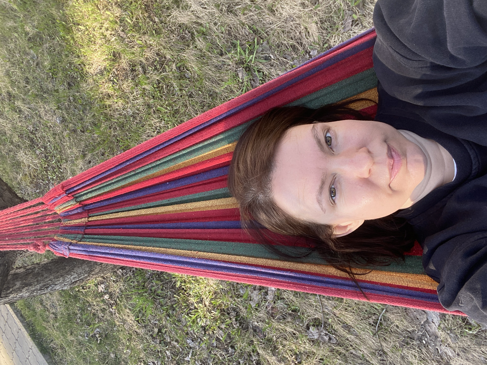 A woman is lying in a colorful striped hammock, suspended between two trees. She is wearing a black top. The ground beneath the hammock consists of grass and dried leaves. The photo appears to have been taken in an outdoor location, possibly a park or backyard. The city name is not evident in the image.