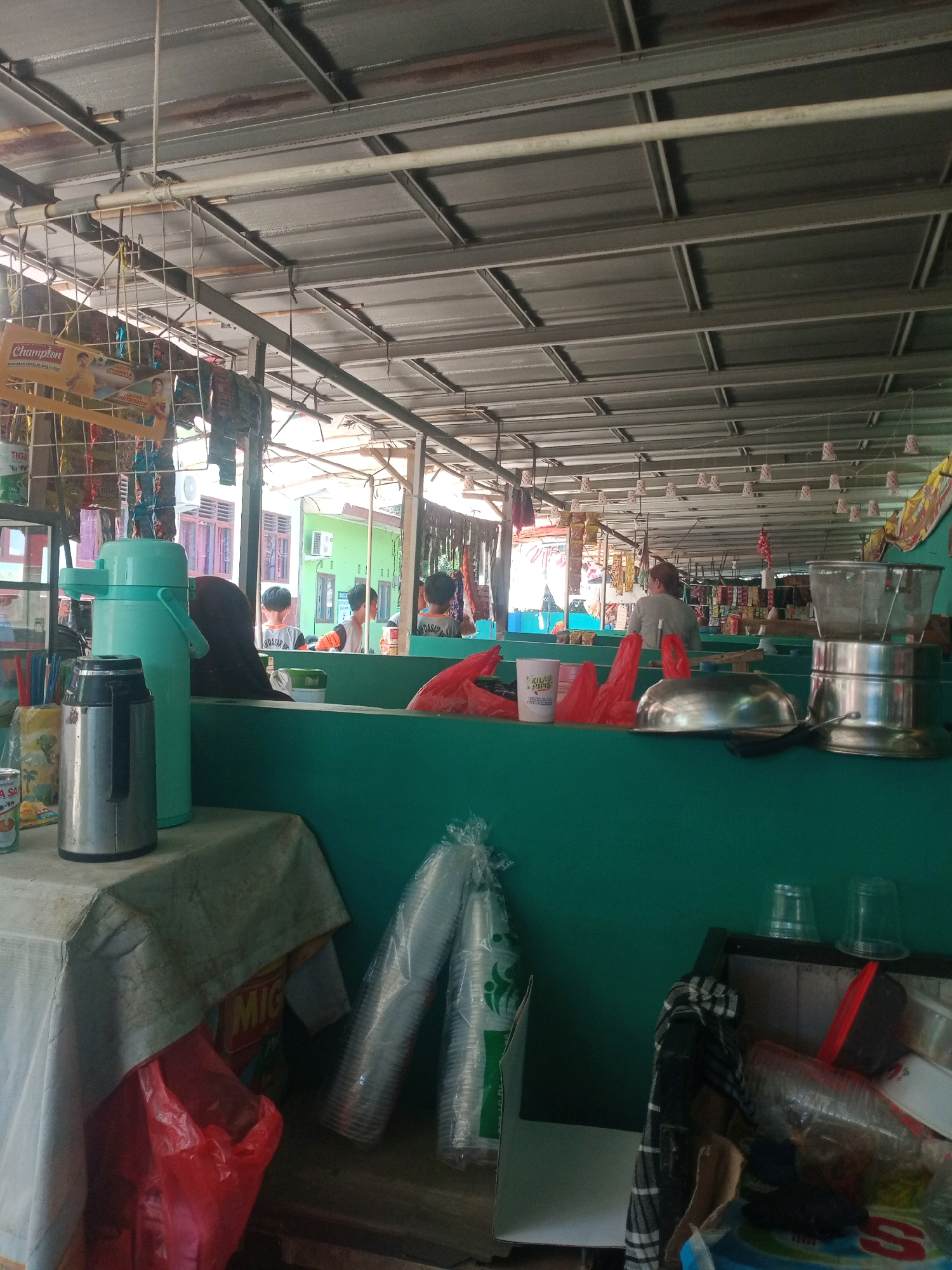 The image depicts an outdoor marketplace stall, possibly a food or drink vendor, under a corrugated metal roof supported by a metal frame. The stall is lined with a vibrant teal-colored counter. In the foreground, a white tablecloth covers a table laden with various items. Prominently displayed are a large silver thermos and a tall, light blue thermos, suggesting beverages are served. A red plastic bag and a stack of plastic cups are also visible on the table.
Behind the counter, several people are seen. Two individuals, appearing to be young males, are facing away from the camera, with their backs to the viewer. Another person, an adult with dark hair and wearing a dark head covering, is partially visible behind the light blue thermos. Further back, more individuals are present, some interacting with the stall or other patrons, and beyond them, the marketplace extends with more stalls and goods displayed. The overall environment suggests a casual, bustling marketplace setting in a city.