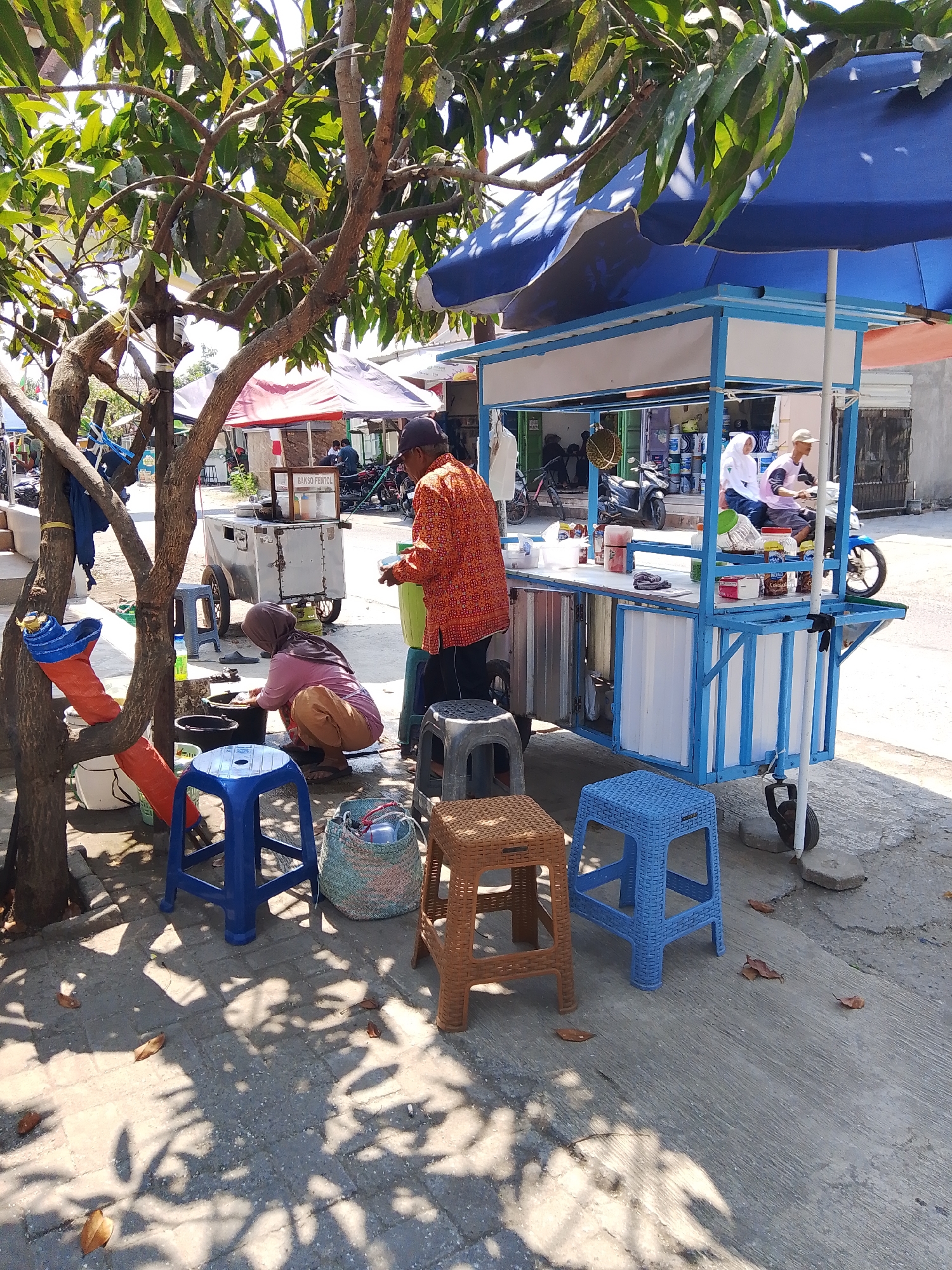 In Kedondong, Indonesia, a street food vendor stands beside a mobile cart under a blue and white awning. The vendor, wearing an orange patterned shirt, appears to be serving a customer. A woman wearing a headscarf and brown pants is crouched nearby, attending to something on the ground. Several plastic stools, a woven basket, and various containers are scattered around the area. A tree with green leaves shades part of the scene. In the background, another food stall with a red awning is visible, as are motorcycles and buildings. The ground is paved with concrete and bricks, with fallen leaves and dappled sunlight creating a textured surface.