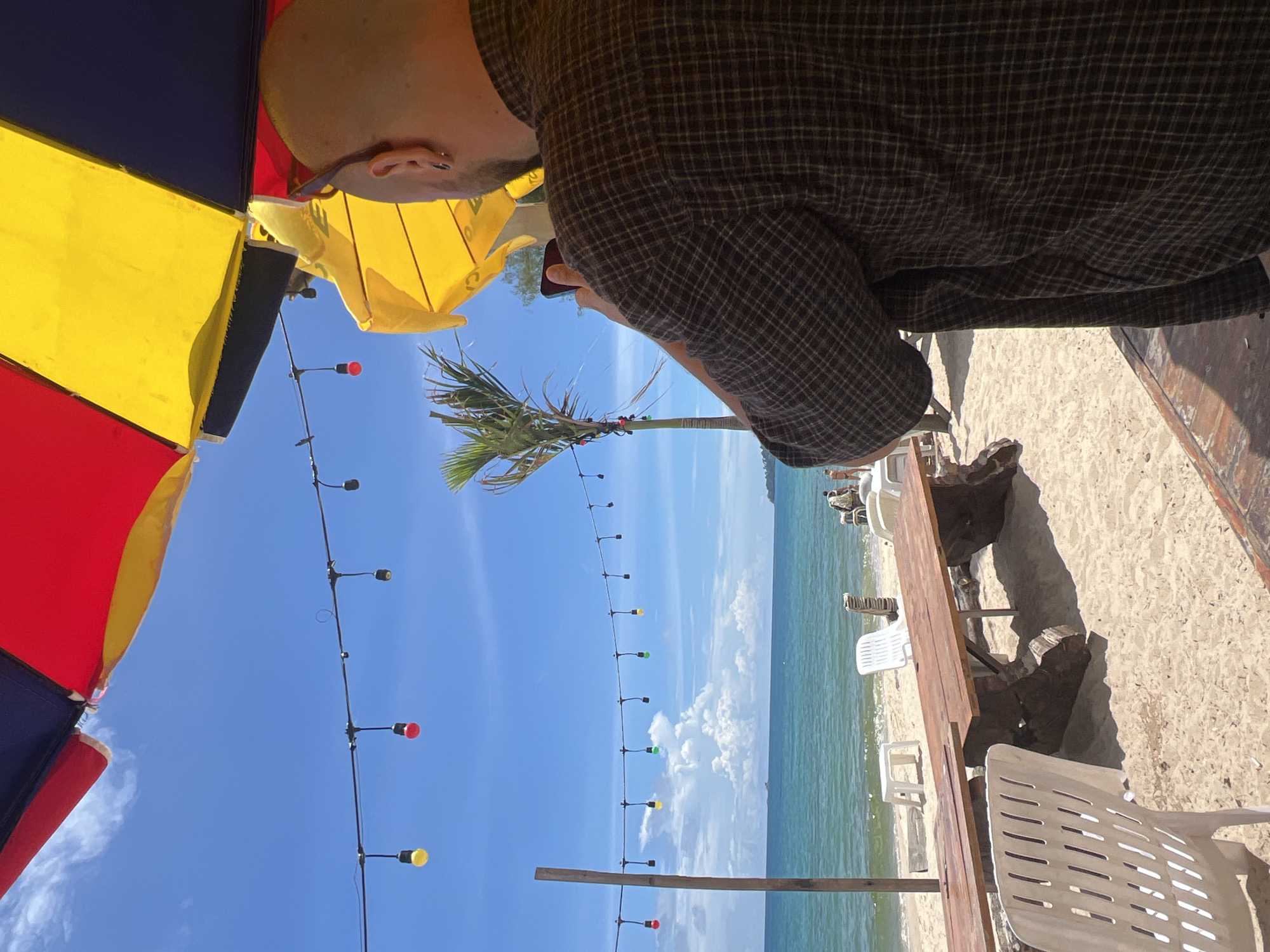 A man wearing a plaid shirt and sunglasses is standing on a sandy beach in Choeng Thale, Thailand, holding a phone up to take a picture. He is facing away from the camera, towards the ocean. In the background, there are several beach chairs, wooden tables, and a palm tree. The sky is clear and blue, with a few scattered clouds. The sun is shining brightly, casting shadows on the sand. There are strings of colorful lights strung across the beach. The ocean is a vibrant turquoise color, and the waves are gentle. In the distance, there are a few small boats on the water.