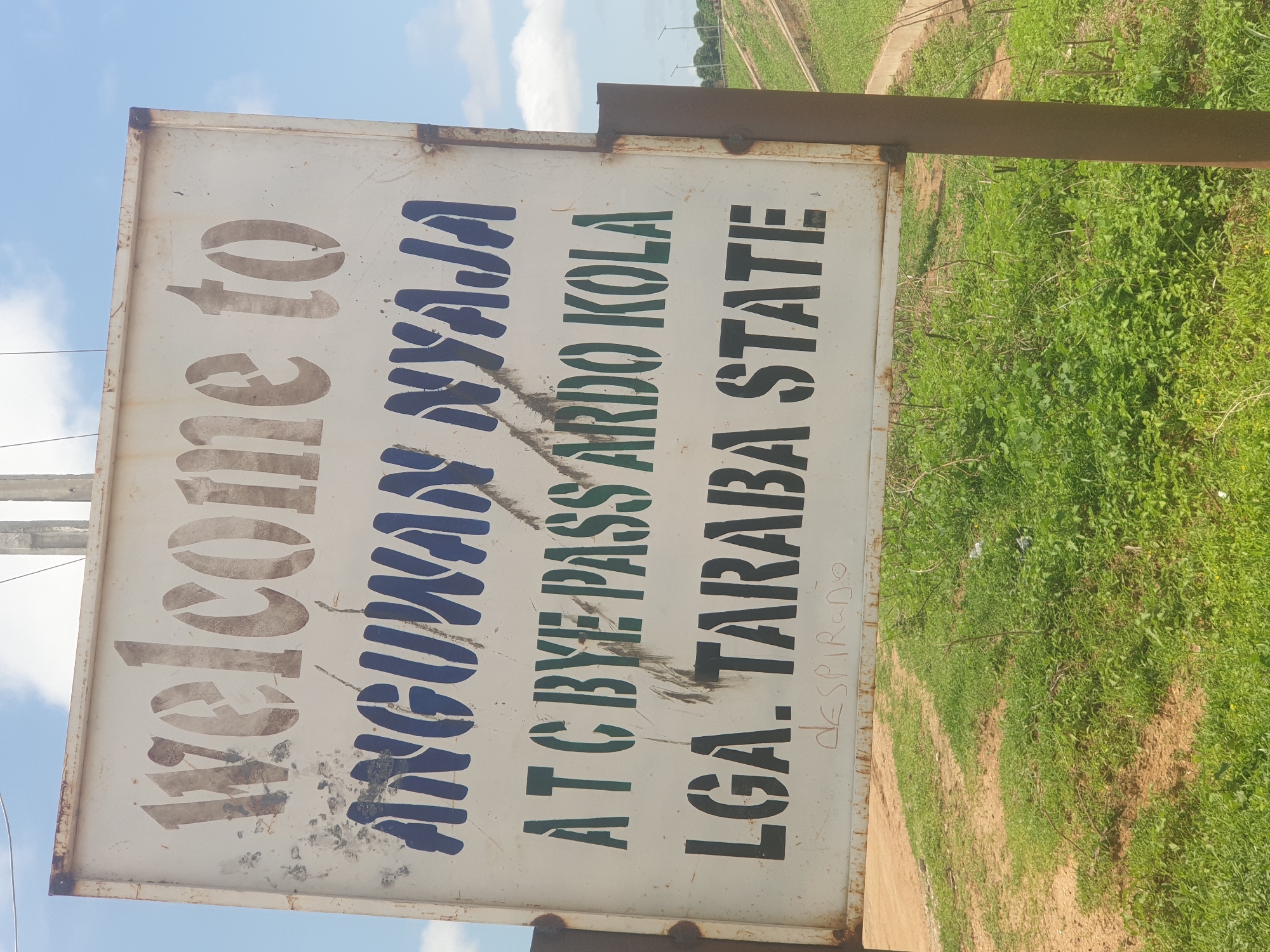 A weathered rectangular welcome sign stands vertically, mounted on two dark metal posts. The sign's white surface is heavily marked with scratches, dirt, and rust, particularly along its edges and central areas. The sky above is clear blue with scattered white clouds.
The sign features text in a stencil font, varying in color. The top line reads "welcome to" in a faded brownish hue. Below it, "ANGUWAN NYAJA" is displayed in blue. The subsequent line, "A T C BYE PASS ARDO KOLA," is in green. The bottom main text, "LGA. TARABA STATE," is in black. Beneath the black text, faint, reddish-brown graffiti spells "dESPIRado".
The text indicates the location as Anguwan Nyaja, situated at the ATC Bye Pass within the Ardo Kola Local Government Area, Taraba State, Nigeria. This places the scene in the vicinity of Jalingo, the capital of Taraba State.
The immediate foreground and midground are dominated by lush green grass and weeds, with a dirt path winding through the vegetation on the left side of the frame. To the right in the background, a gently sloping landscape is visible, possibly agricultural land or a cleared area, marked by several distant light poles.