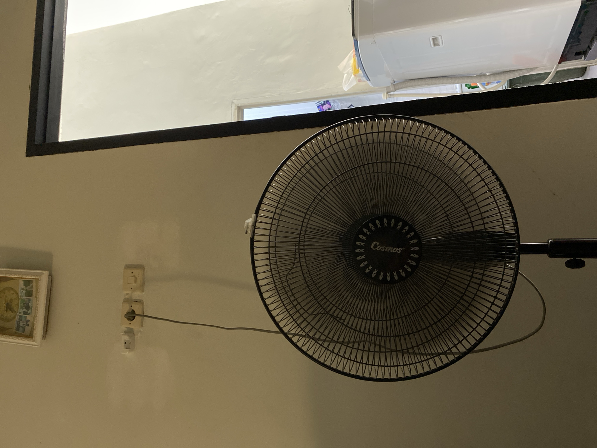 The image depicts an indoor scene with a floor fan positioned centrally. The fan is dark-colored with a visible brand name "Cosmos" on its grille. To the left of the fan, a wall is visible, adorned with a framed picture at the top and two electrical outlets with a plugged-in cord. To the right, a doorway leads to another area where a white washing machine is partially visible. The lighting suggests it is daytime, with light coming in from the doorway. The location is stated to be in Babelan, Indonesia.