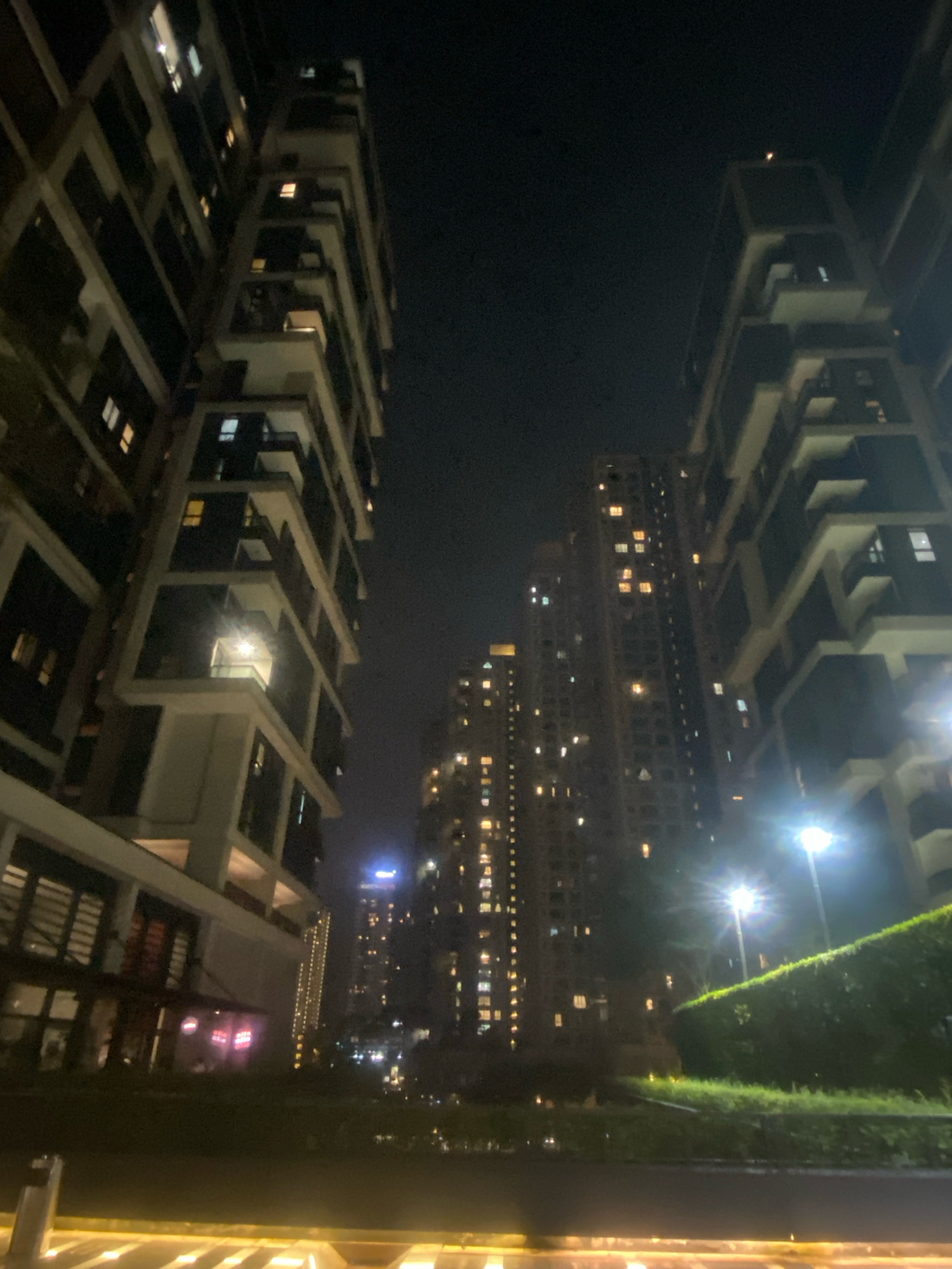 The image depicts a nighttime scene in Kuala Lumpur, Malaysia, focusing on the modern architecture of high-rise apartment buildings. The perspective is low-angle, looking up at the structures that dominate the frame. The buildings are illuminated by numerous lit windows, suggesting occupancy. One building on the left, closer to the viewer, has a more detailed facade with balconies and dark-paned windows. To the right, another taller building displays a grid of glowing windows stretching upwards. In the mid-ground, a cluster of even taller buildings with a dense pattern of lights can be seen. A streetlamp with a bright light source is visible in the right foreground, casting a glare. Lush green hedges and grass areas are situated in front of the buildings, illuminated by the artificial lights. The sky is dark, indicative of night. The overall impression is one of an urban residential environment characterized by tall structures and artificial lighting.
