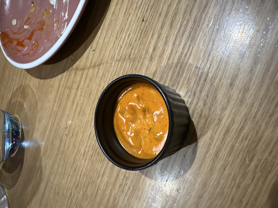 A small, black ceramic fluted ramekin bowl, approximately 7-8 cm in diameter, containing an orange-red creamy sauce, is centrally positioned on a light brown wooden table surface. The sauce exhibits a textured appearance with darker red or brown flecks, consistent with herbs or spices. In the upper left quadrant, a partial view of a clear glass filled with a translucent light blue liquid is visible. To the upper right, a segment of a round ceramic plate with a distinct pink rim and a white underside is present. This plate displays remnants of reddish sauce and small food particles, indicating recent use. Shadows are cast to the lower right of the ramekin and below the glass, suggesting an overhead light source. The scene depicts a dining context, likely within a restaurant or eatery located in Kalyan-Dombivli, India. The precise address or specific establishment within Kalyan-Dombivli cannot be determined from the visual evidence provided.