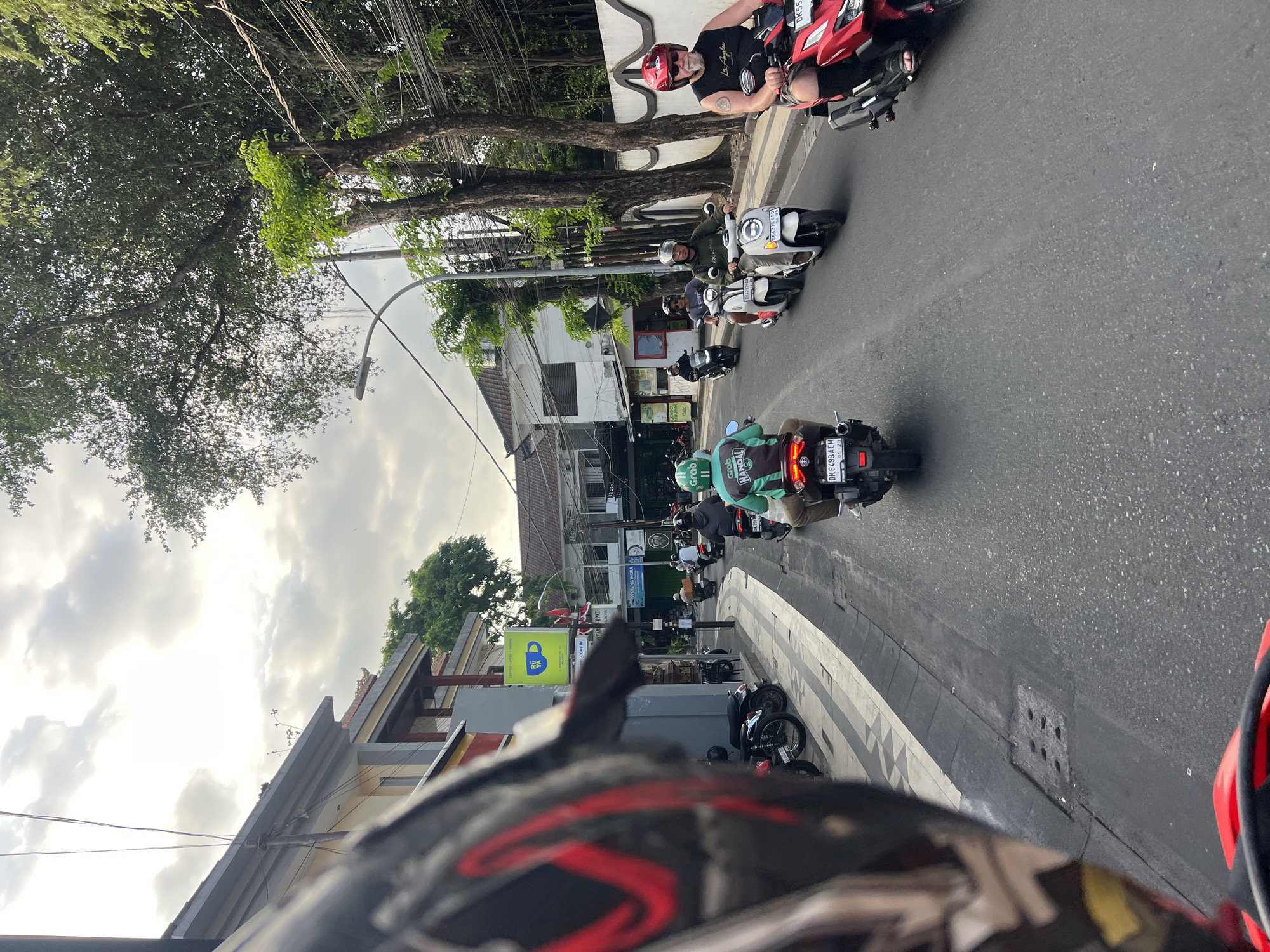 The image depicts a street scene in Kuta, Indonesia, likely during daytime with overcast skies. The perspective is from a rider on a motorcycle, with the rider's helmet dominating the foreground. The street is lined with buildings on the left, featuring a mix of modern and traditional architecture, and lush trees on the right. Several people are riding motorcycles and scooters, moving in the same direction. On the left side of the street, there are parked motorcycles and a building with a sign for "RUYA COFFEE". The road surface is asphalt. The overall atmosphere suggests a busy urban environment with significant motorcycle traffic.