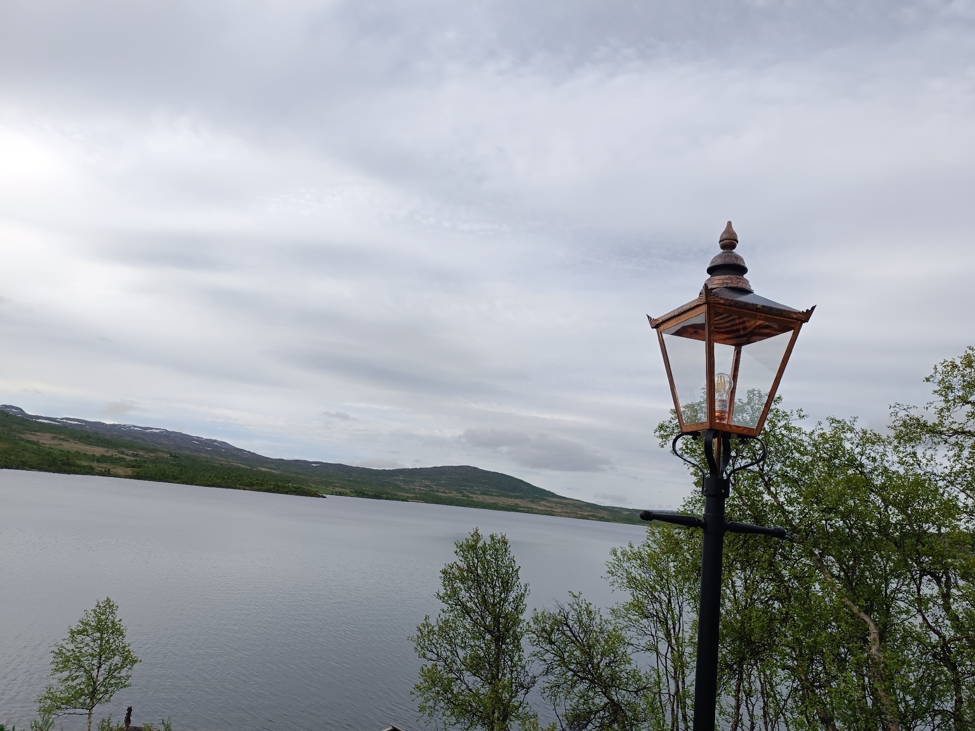 A classic outdoor lantern with a copper-toned roof and glass panels stands to the right of the frame, attached to a black metal pole. The lantern's bulb is visible. In the background, a large body of calm, gray water stretches across the image, bordered by a lush green, hilly landscape that recedes into the distance. The sky is overcast with a soft, diffused light. The scene appears to be located in Hovden, Norway, suggesting a natural, possibly remote, setting.