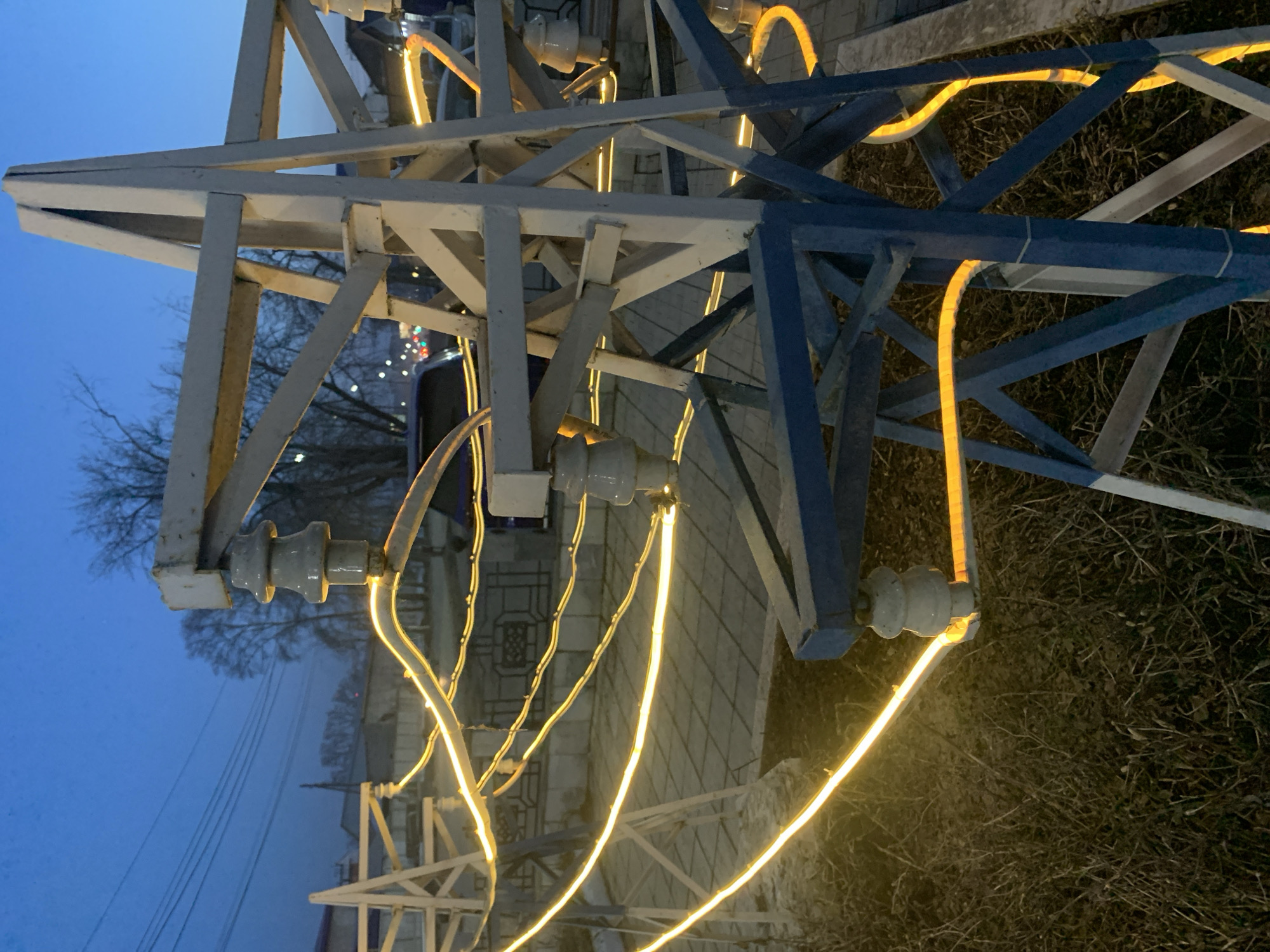 The image shows an electrical power transmission tower in Chaikovsky, Russia. The tower is constructed of steel, with some parts painted white and others blue. Electrical insulators are visible, supporting what appear to be power cables or wires. The tower is decorated with yellow rope lights. The background includes a building, trees without leaves, and a blue sky. The ground around the tower is covered with dry vegetation.