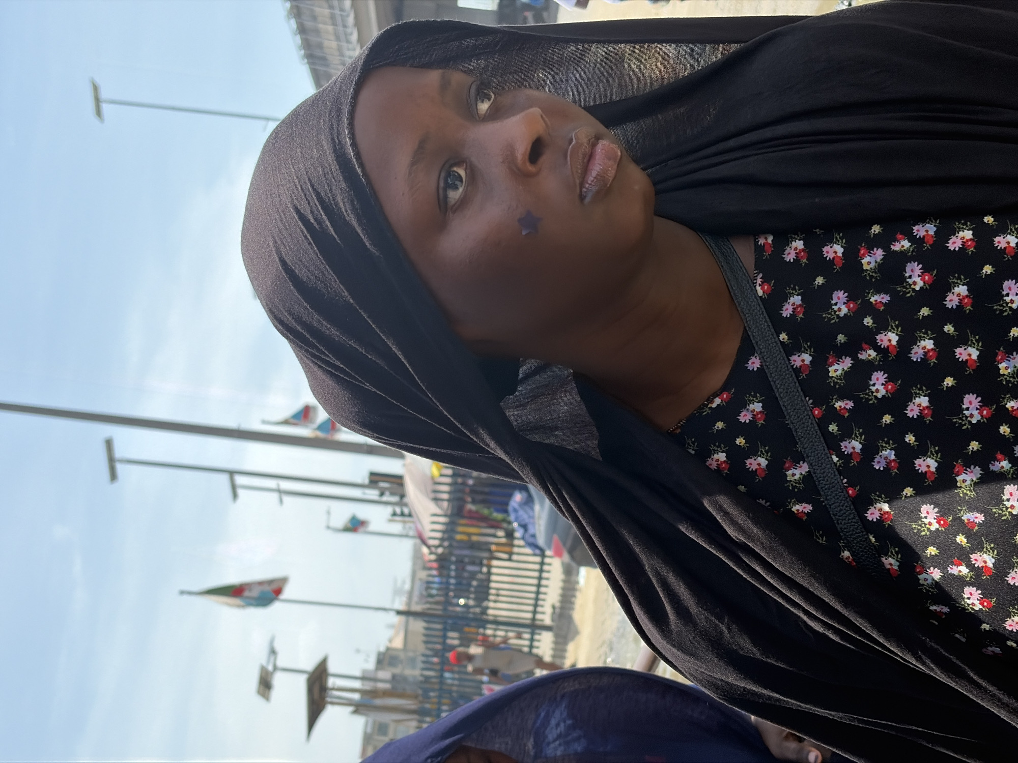 A woman wearing a black headscarf and a black floral-patterned top stands on the side of a street in Lekki, Nigeria. A small blue star is affixed to her cheek. The location is crowded. Light poles displaying flags are visible along the street. A person is visible near a blue metal fence behind her. Other indistinct figures are in the background. The sky is light blue.