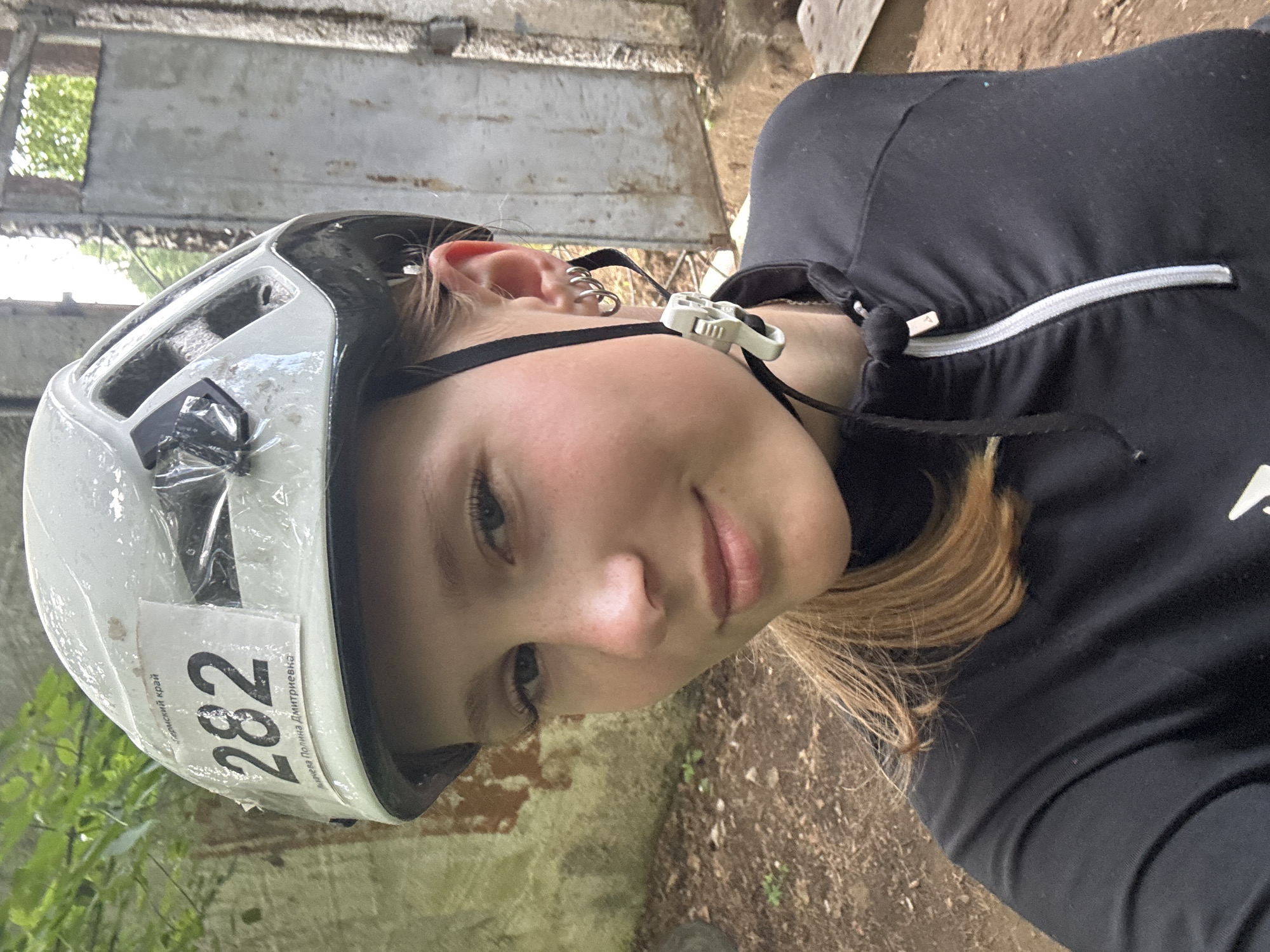 A young woman with blonde hair, wearing a black zip-up jacket and a white helmet with the number 282, takes a selfie outdoors. The helmet has a label with Russian text and the number 282. She is also wearing hoop earrings. The background shows a dirt path, some greenery, and a weathered metal gate or structure. The scene appears to be in a park or outdoor recreational area.