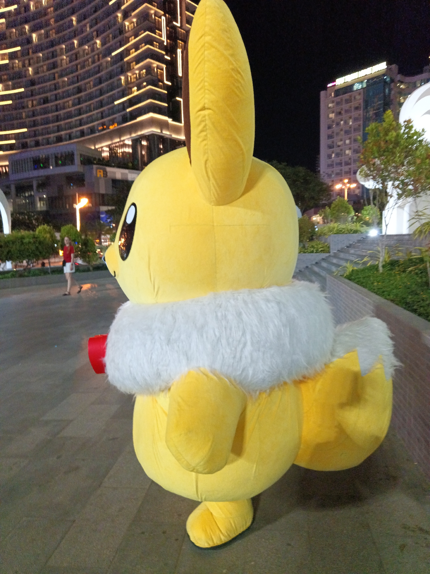 An Eevee mascot costume, a large yellow plush character with a white fur collar, stands in the foreground of the image. The mascot is seen from the side, facing left, with its large ear pointing upwards. A person wearing a red shirt and white shorts is visible walking in the background, to the left of the mascot. The background features a curved, modern building with many illuminated windows, indicating an urban setting at night. The ground is paved with square tiles. The scene is lit by artificial lights from the buildings and streetlights. This image was taken at Nha Trang Ward, Vietnam.
