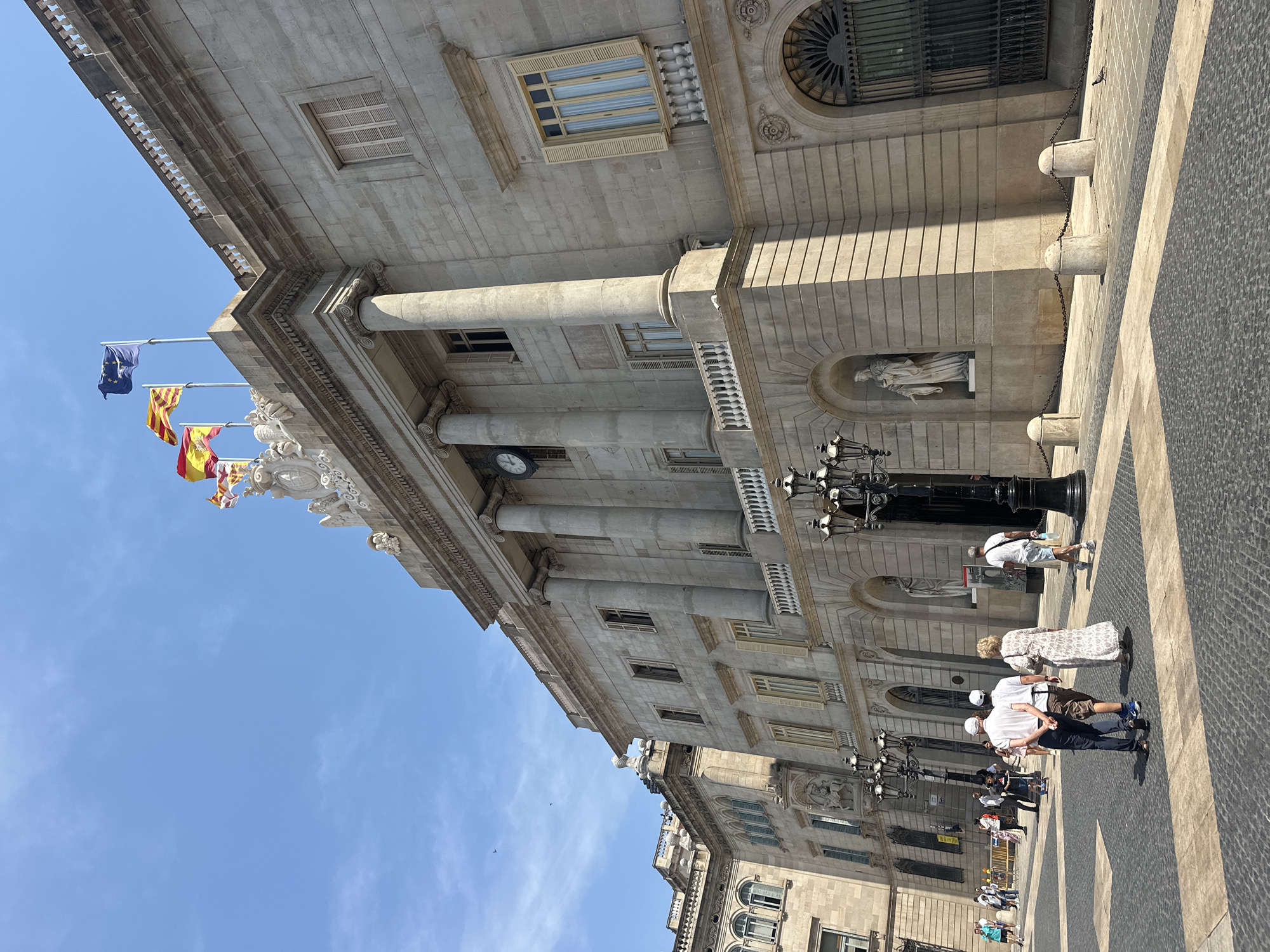 The image captures a view of the Ajuntament de Barcelona (City Hall of Barcelona), Spain, from the Plaça de Sant Jaume. The building is constructed of light-colored stone and features a classical architectural style with columns, arched windows, and decorative elements. Three flags are visible flying from the rooftop, one of which is the European Union flag, and another appears to be the flag of Spain. In the foreground, several people are walking across a paved plaza. The plaza is designed with contrasting patterns of dark and light stones. Several ornate street lamps are also present. The sky is clear and blue with a few wispy clouds. The scene is bathed in bright sunlight, casting distinct shadows.