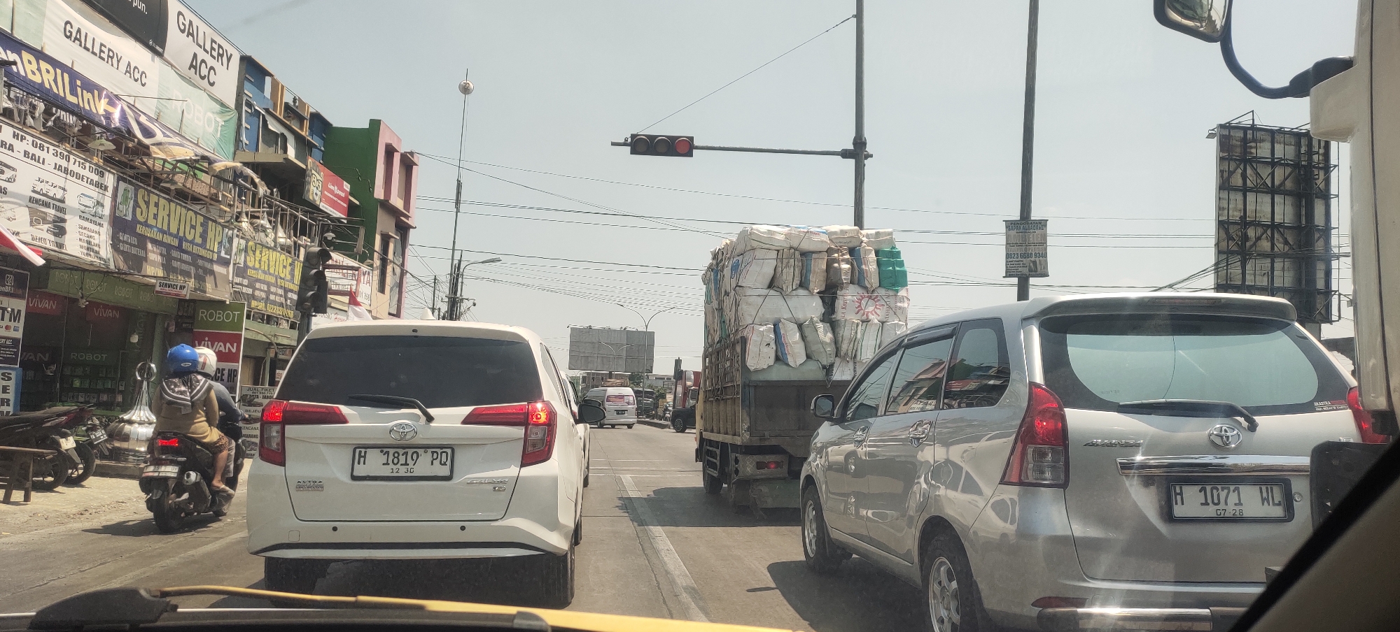 The image depicts a street scene in Sedo Lor, Indonesia, characterized by moderate traffic under a clear, bright sky. The perspective is from inside a vehicle, looking forward.
In the foreground, the rear of a white Toyota Avanza is visible, with its license plate reading "H 1819 PQ" and "12 3C". To the right of the white Avanza, a silver Toyota Avanza is also in view, its license plate displaying "H 1071 WL" and "07 28".
A motorcycle carrying two individuals, a driver and a passenger, travels ahead in the left lane. The driver is wearing a blue helmet, and the passenger has a backpack. The motorcycle has its license plate partially obscured.
Further ahead, a large truck loaded with numerous sacks is prominent in the right lane. The sacks appear to be secured with netting. Traffic lights are visible, with the red light illuminated. Various buildings line the street, displaying multiple signs with text, some of which indicate commercial establishments like "GALLERY ACC", "SERVICE HP", and "VIVAN". Billboards and utility poles are also present. The road surface is asphalt. The overall environment suggests an urban or semi-urban setting.