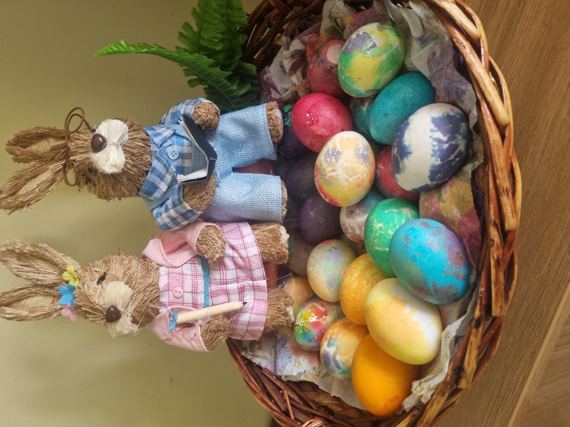 A basket of Easter eggs and two bunny figurines sit on a surface in Unknown, Unknown. The basket, constructed of woven brown reeds, is brimming with vibrantly colored Easter eggs in shades of red, yellow, green, blue, orange, and purple, arranged on a base of crumpled, patterned tissue paper. Beside the basket, two straw bunny figurines lie horizontally. The top bunny wears a blue plaid shirt and glasses, while the bottom bunny sports a pink plaid shirt and holds a pencil. A green fern sits between the basket and the top bunny. The surface on which the items rest is light-colored.