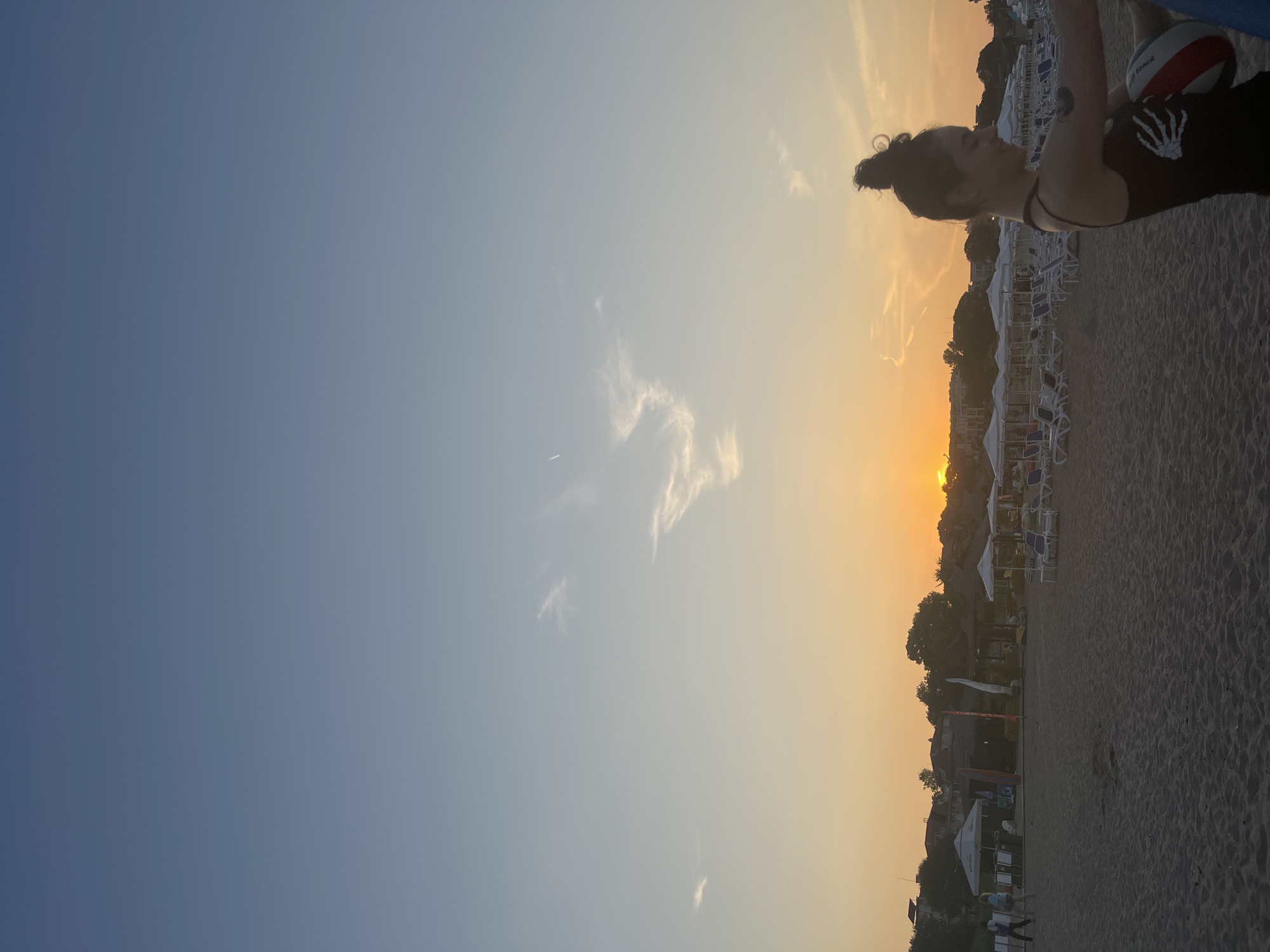 The image depicts a sunset scene on a sandy beach in Lozenets, Bulgaria. The sky transitions from a deep blue at the top to a warm orange near the horizon where the sun is setting. Wispy clouds are visible in the sky. In the foreground, a young woman with her hair in a bun is silhouetted against the sunset. She is wearing a black tank top with a white skeleton hand graphic on it and is holding a beach ball. Behind her, the beach is lined with rows of empty lounge chairs and umbrellas, suggesting it is late in the day. In the middle ground, there are some buildings and trees visible, and a few people can be seen walking on the sand. The overall atmosphere is peaceful and serene, capturing the end of a day at the beach.