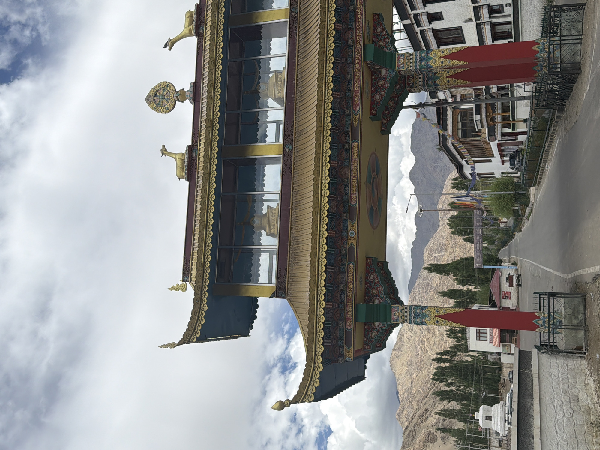 A wide-angle, eye-level outdoor shot captures a Buddhist stupa in Thiksey, India, under a partly cloudy sky. The stupa is a white, dome-shaped structure with a spire, located on the left side of the frame. In the foreground, a road runs through the center, flanked by buildings on both sides. On the right, traditional Tibetan-style architecture is visible, with white walls and dark window frames. On the left, a small white building with a red awning is present. The background is dominated by barren, mountainous terrain, with a sparse scattering of tall, green trees lining the road. A vibrantly decorated gateway, a prominent feature of the scene, stands in the middle ground, adorned with gold accents, colorful paintings, and intricate carvings. Two golden deer statues and a Dharma wheel symbol are visible on the roof of the gateway. The overall atmosphere is serene and spiritual, with a clear focus on the architectural elements and the natural landscape.
