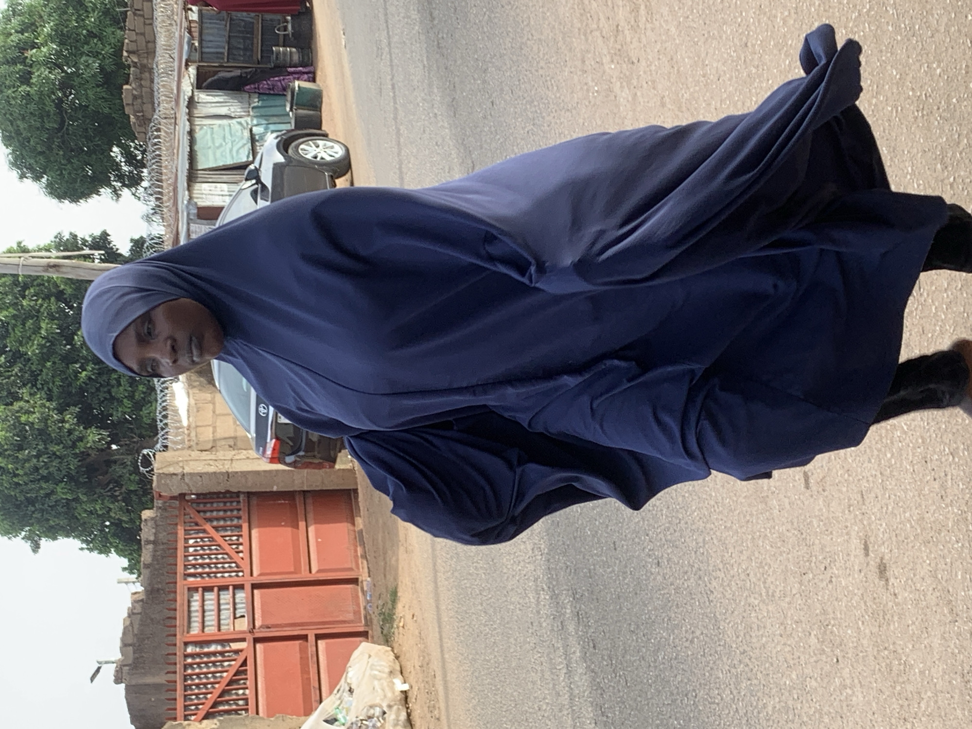 A person wearing a dark blue full-body covering walks along a road in Jos, Nigeria. In the background, there is a gated fence, and parked vehicles. Other buildings line the street, with trees in the vicinity.
