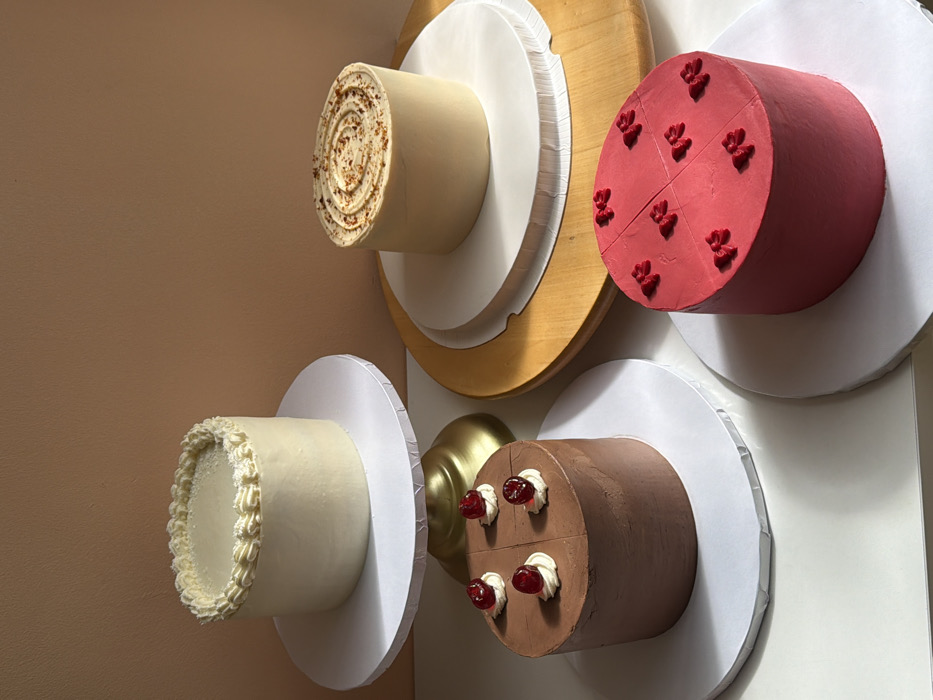 The scene displays four individual, decorated cakes arranged on a white surface against a plain, light brown wall in an indoor setting. The specific location within Tel-Aviv, Israel, is not discernible from the visual information.
A cylindrical cake, covered in off-white frosting with a piped scalloped border and textured topping, is positioned on a white cake stand with a golden pedestal base on the left. To its right, another cylindrical cake, light tan in color with a spiral crumb design on its top, rests on a white cake board placed on a light brown wooden turntable.
In the foreground, a dark brown cylindrical cake with its top divided into four quadrants, each featuring a dollop of white frosting topped with a red glacé cherry, sits on a white cake board. Adjacent to it on the right is a bright red cylindrical cake, also with a quartered top, adorned with eight small red bow-shaped decorations.
The white surface supports the cakes directly or via boards and stands. Illumination from the upper left casts distinct shadows on the white surface. No people, actions, or interactions are present.