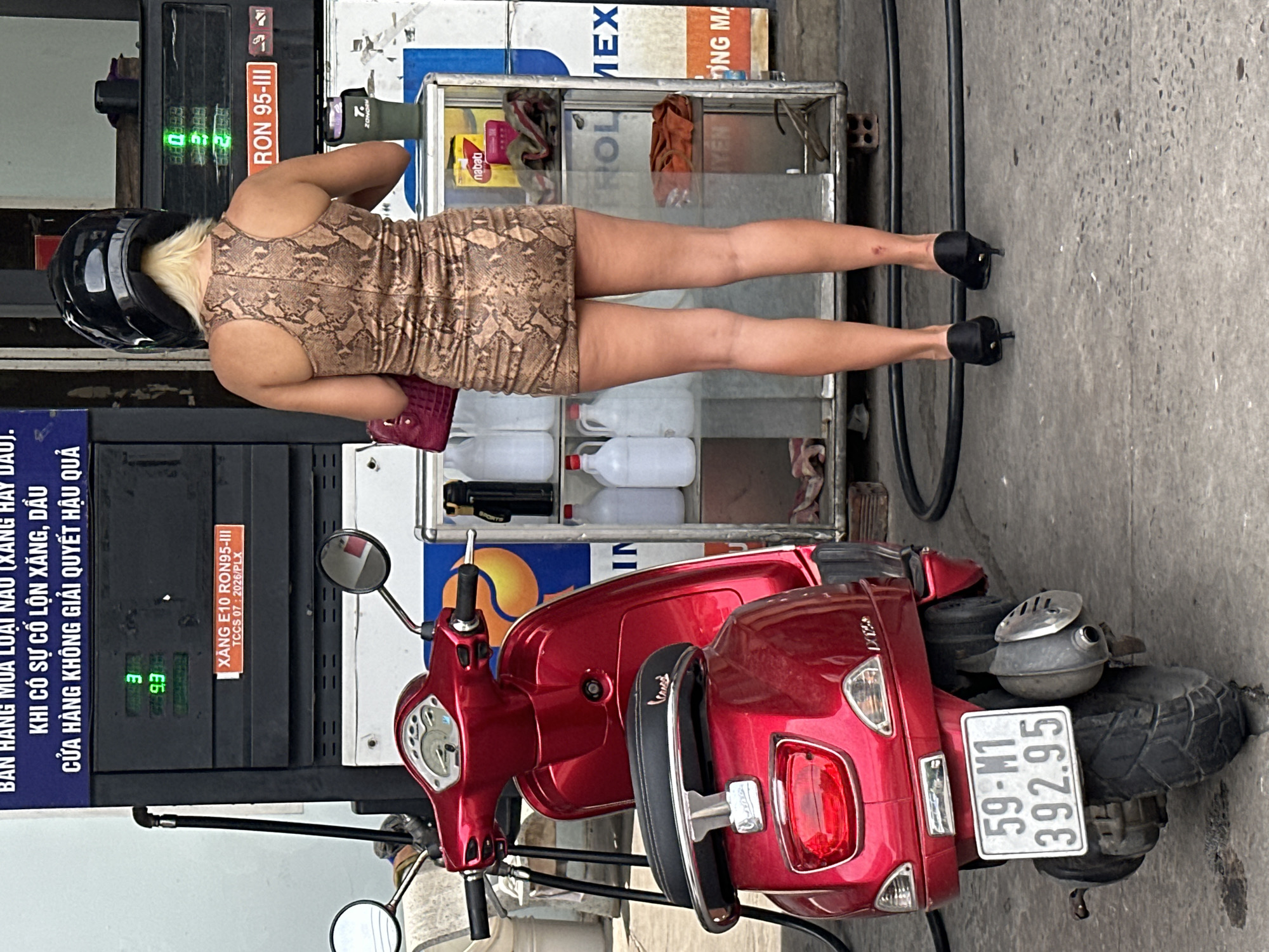 A woman wearing a black helmet and a short, snake-print dress stands at a gas station in Bac Nha Trang Ward, Vietnam. Her blonde hair is visible beneath the helmet. She is wearing black high heels and holding a red purse. A red scooter is parked to her left. Behind her, a gas pump displays prices and product information. A glass display case containing various items, including bottles and packaged goods, is visible to her right. The scene is outdoors on a paved surface, with a wall and more gas pumps in the background.