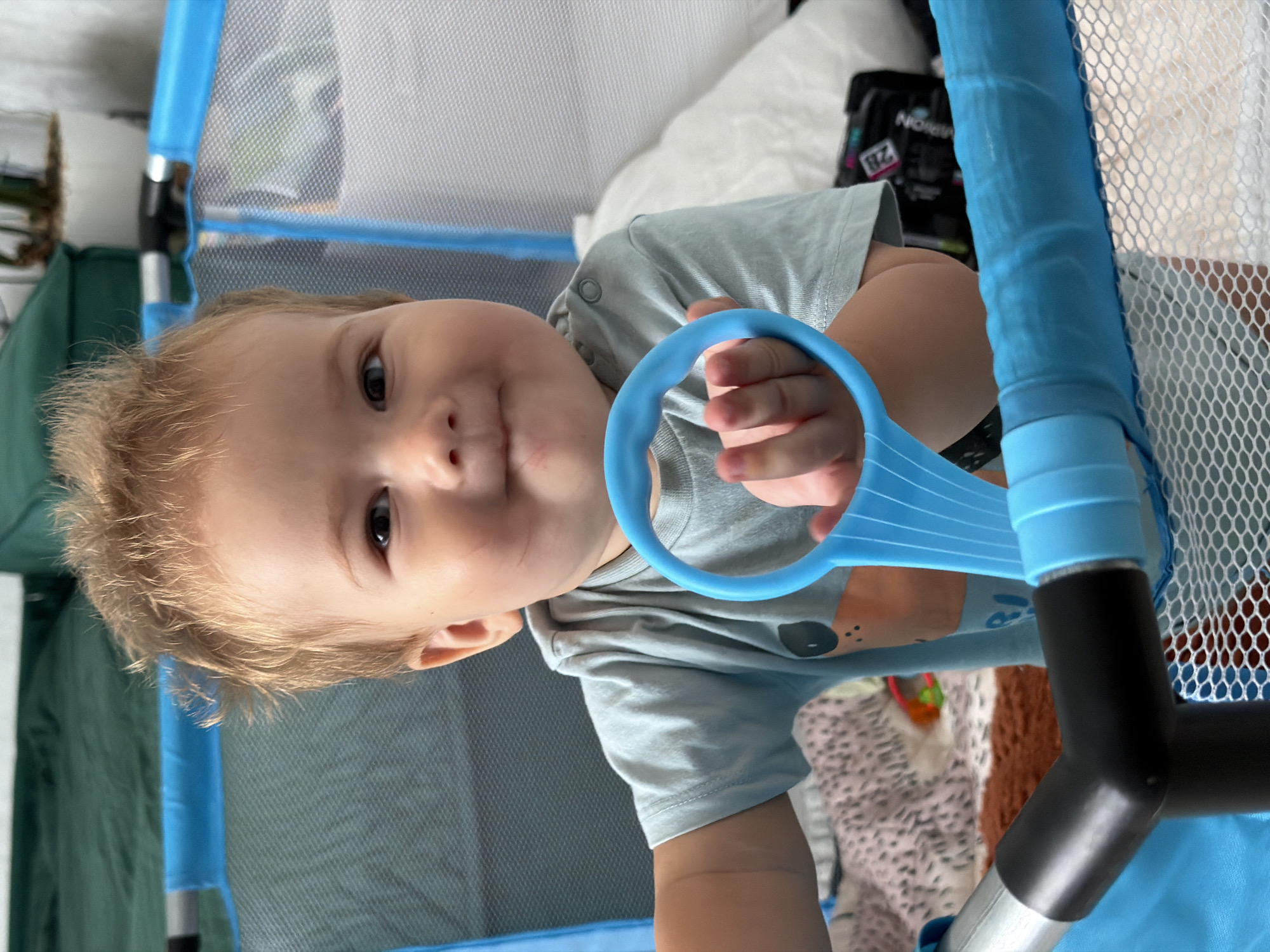 A young child with blond hair is inside a blue playpen. The child is wearing a light blue t-shirt with a graphic design. The child is holding a blue toy with a circular handle. The playpen is made of blue plastic tubing and mesh netting. In the background, there is a green sofa and a white pot with a plant. The scene appears to be indoors, possibly in a living room. The image does not contain any information to determine the location in the city of Kazan, Russia.
