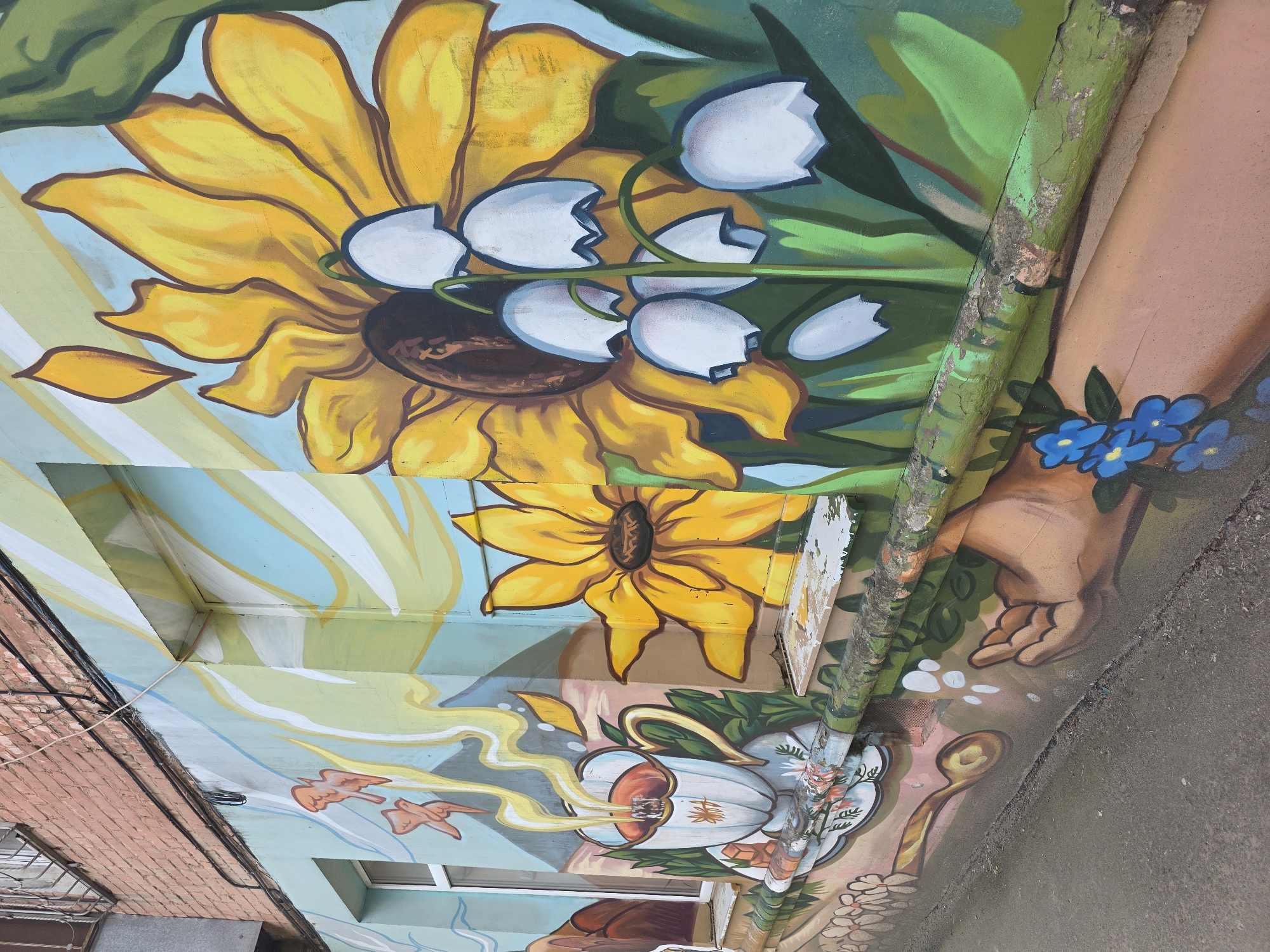 The image shows a section of wall art in Krasnodar, Russia, specifically on a building at st. Rashpilevskaya, 145. The mural features a sunflower and lilies of the valley at the top. Below, there is a teacup depicted with steam in the form of birds, and a spoon painted alongside. A hand is also part of the mural, with small blue flowers near the wrist. A green rain pipe runs along the wall. The background is a mixture of sky-blue, beige, and green tones.