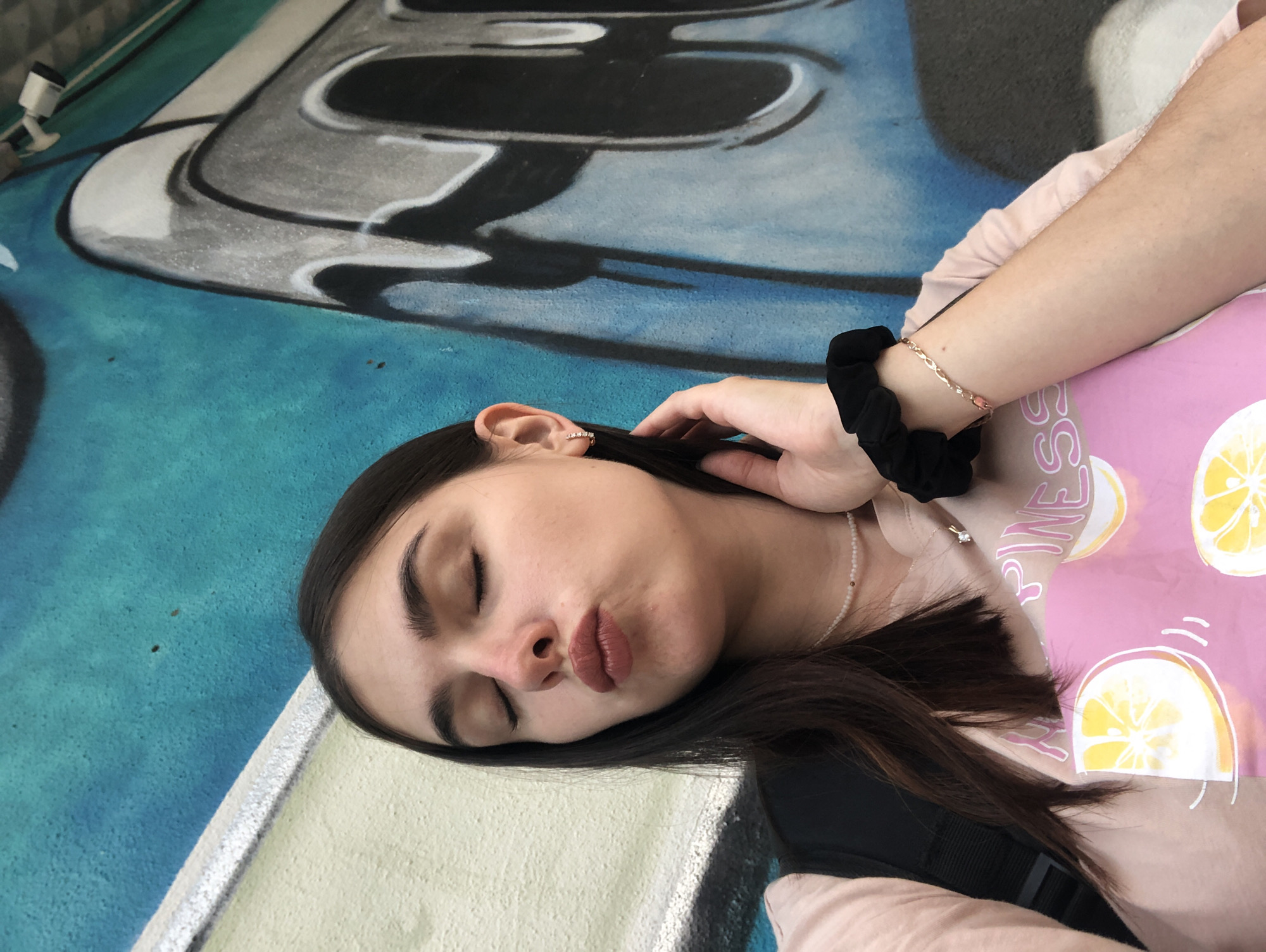 A young woman with dark hair is lying down with her eyes closed, appearing to be asleep or resting. She is wearing a light pink t-shirt with a pattern of sliced lemons. A black scrunchie is visible on her left wrist, and she wears a delicate necklace with small pearls and a single pendant. Her right hand is resting on her head, touching her hair. The background features a colorful mural with abstract shapes and designs in blue, gray, and white. A small white security camera is mounted on the wall in the upper left corner of the image. The lighting suggests it is daytime, and the overall atmosphere is casual and relaxed. The image appears to be a self-portrait taken from a low angle. No text is clearly visible in the image.