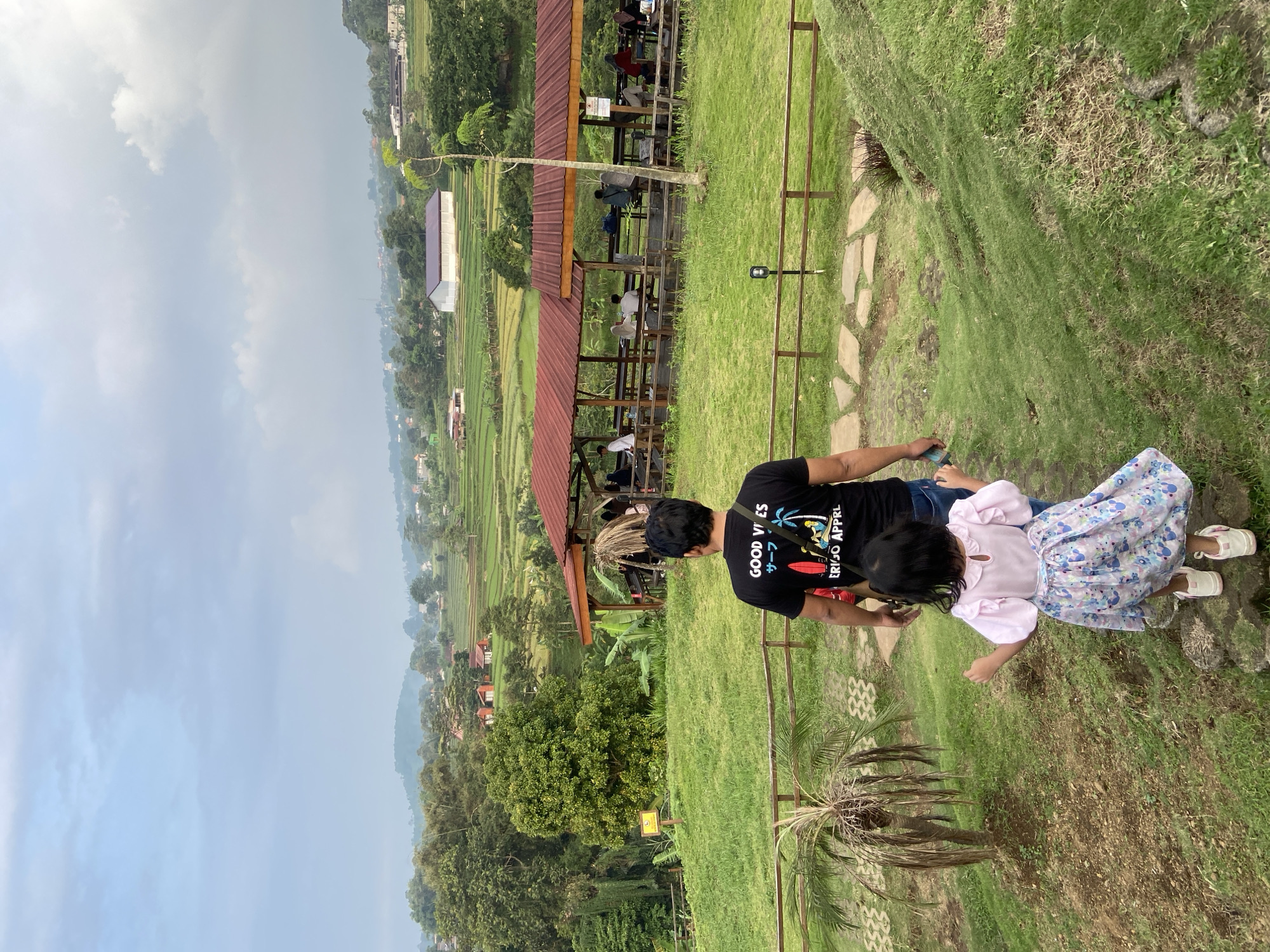 The image captures a man and a young girl standing on a grassy hillside, overlooking a landscape. The scene is located in Tamiajeng, Indonesia. The man is wearing a black t-shirt with the words "GOOD VIBES" and "ERIGO APPRL" printed on it. The girl is wearing a pink top and a dress with a floral pattern. In the background, there is a wooden structure with people seated inside. Beyond the structure, there are tiered rice paddies and houses, stretching into the distance. The sky is overcast with clouds.