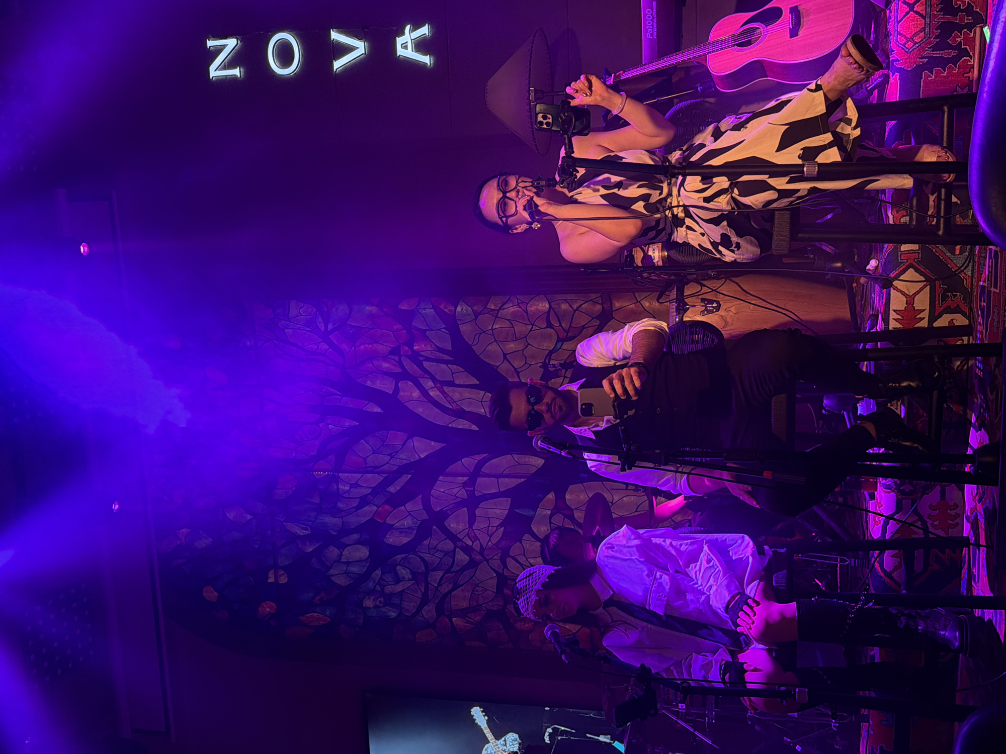 In Yuzhong District, China, on a dimly lit stage bathed in purple light, a band performs. A female singer, wearing a black and white patterned dress, stands at a microphone, her hand raised to her mouth. A man in sunglasses and a vest holds up his phone, possibly recording the performance. Another band member, wearing a white shirt and a headscarf, stands near a drum set. A pink acoustic guitar leans against a speaker. The backdrop features a stained-glass-like design of a tree with branches and leaves. Neon letters spelling "NOVA" glow on the wall behind the performers. The scene suggests a live music event in a venue.