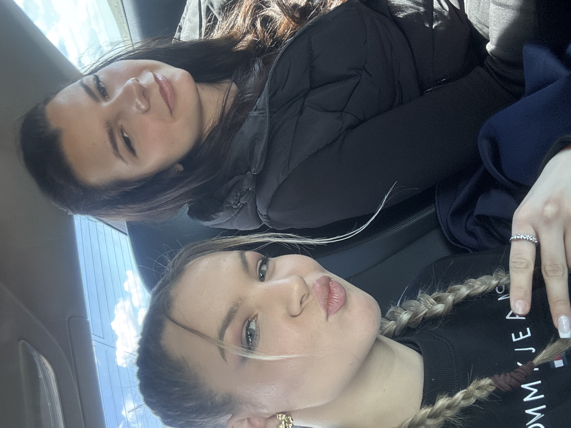 Two young women are in what appears to be a car. The woman in the foreground has blonde hair in braids, pinkish makeup, and is wearing a dark sweatshirt with a Tommy Jeans logo. The woman in the background has dark hair and is wearing a black puffer jacket. The image seems to have been taken from a low angle, looking slightly up at the subjects. The location is not discernible as being in Vratitsa, Bulgaria, or any other specific location, based on visual information. The only environmental detail visible is a portion of a car window with a view of the sky.