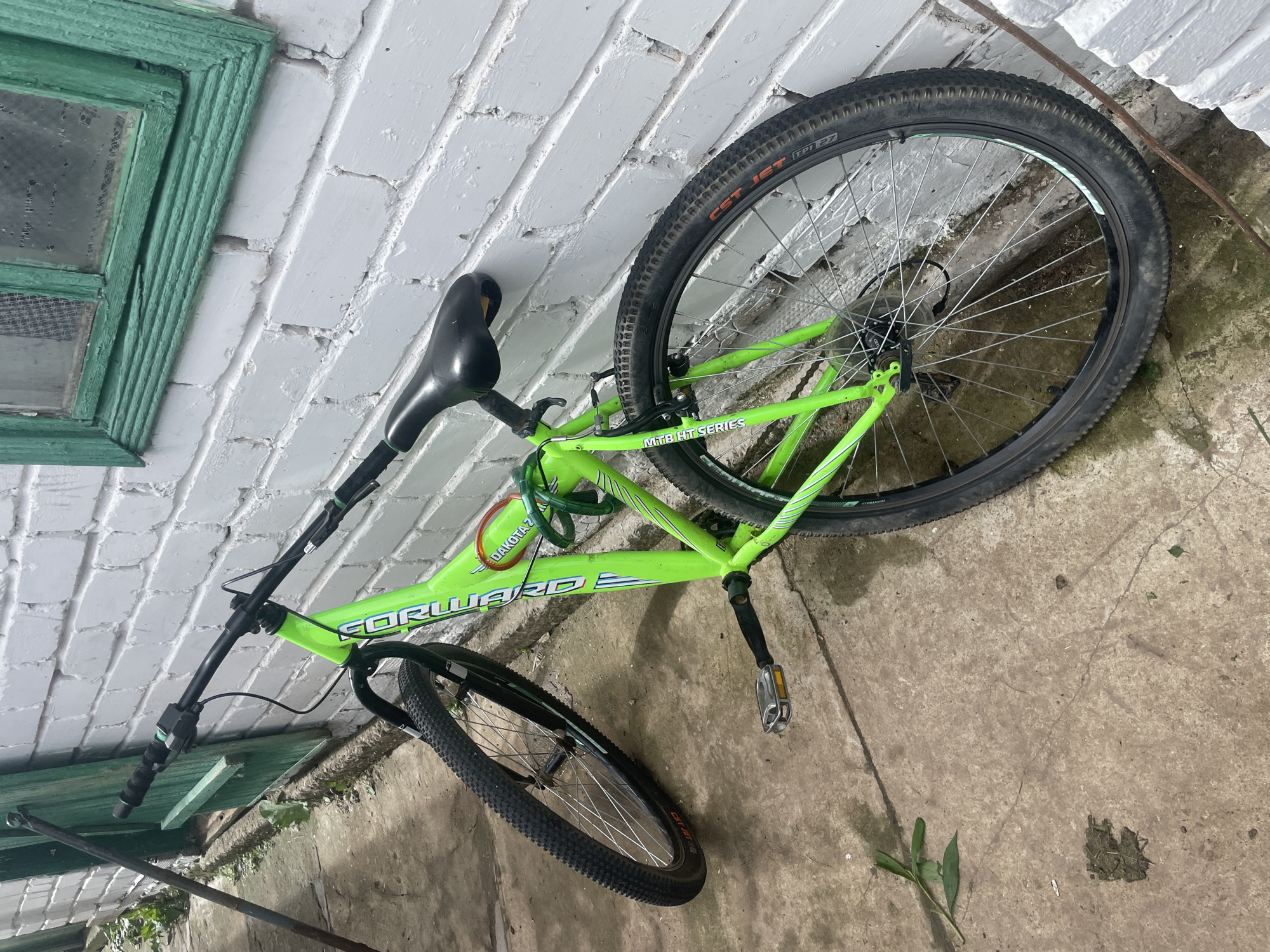 The image shows a bright green bicycle leaning against a white brick wall. The bicycle is positioned in a relaxed manner, with its front wheel slightly turned and resting against the wall. The bicycle has a black seat, black handlebars with grips, and two black tires with silver spokes. A green cable lock is wrapped around the frame and seat post. The bicycle appears to be outdoors, with a concrete ground surface visible in the foreground, scattered with a few leaves and debris. A green wooden window frame with a visibly aged and somewhat peeling paint is present on the wall behind the bicycle. A dark metal pole leans against the left side of the wall, adjacent to a green wooden structure that appears to be a door or gate. The overall environment suggests a residential or utilitarian outdoor area. No people are present in the image. The location is identified as Laishevo, Russia.