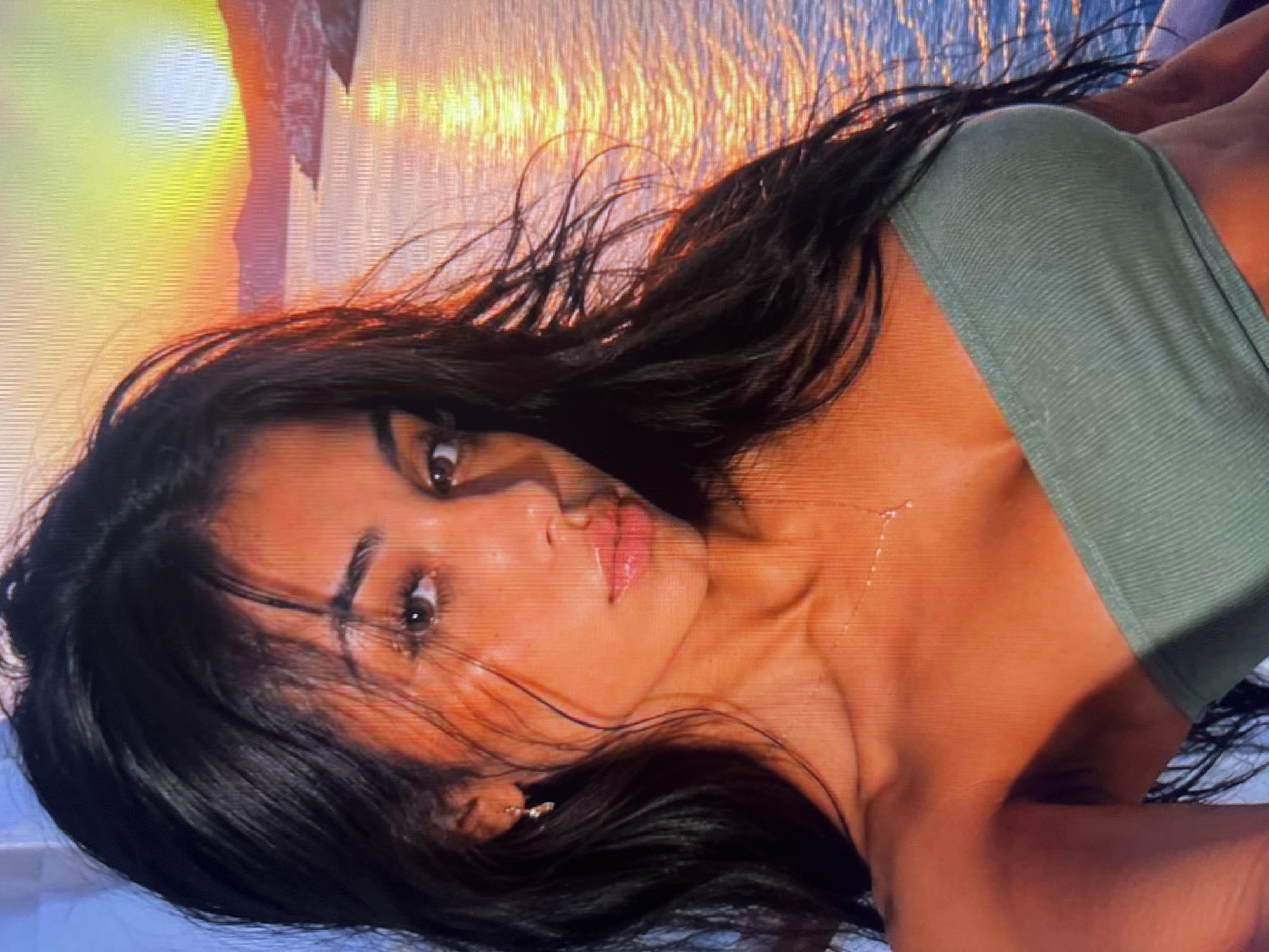 The image is a close-up portrait of a young woman with long, dark hair. She is wearing a green strapless top and a delicate necklace. Her skin is bronzed, and she has makeup on. In the background, a sunset is visible over a body of water with a coastal town or village on the hills. The lighting suggests it is either early morning or late evening.
