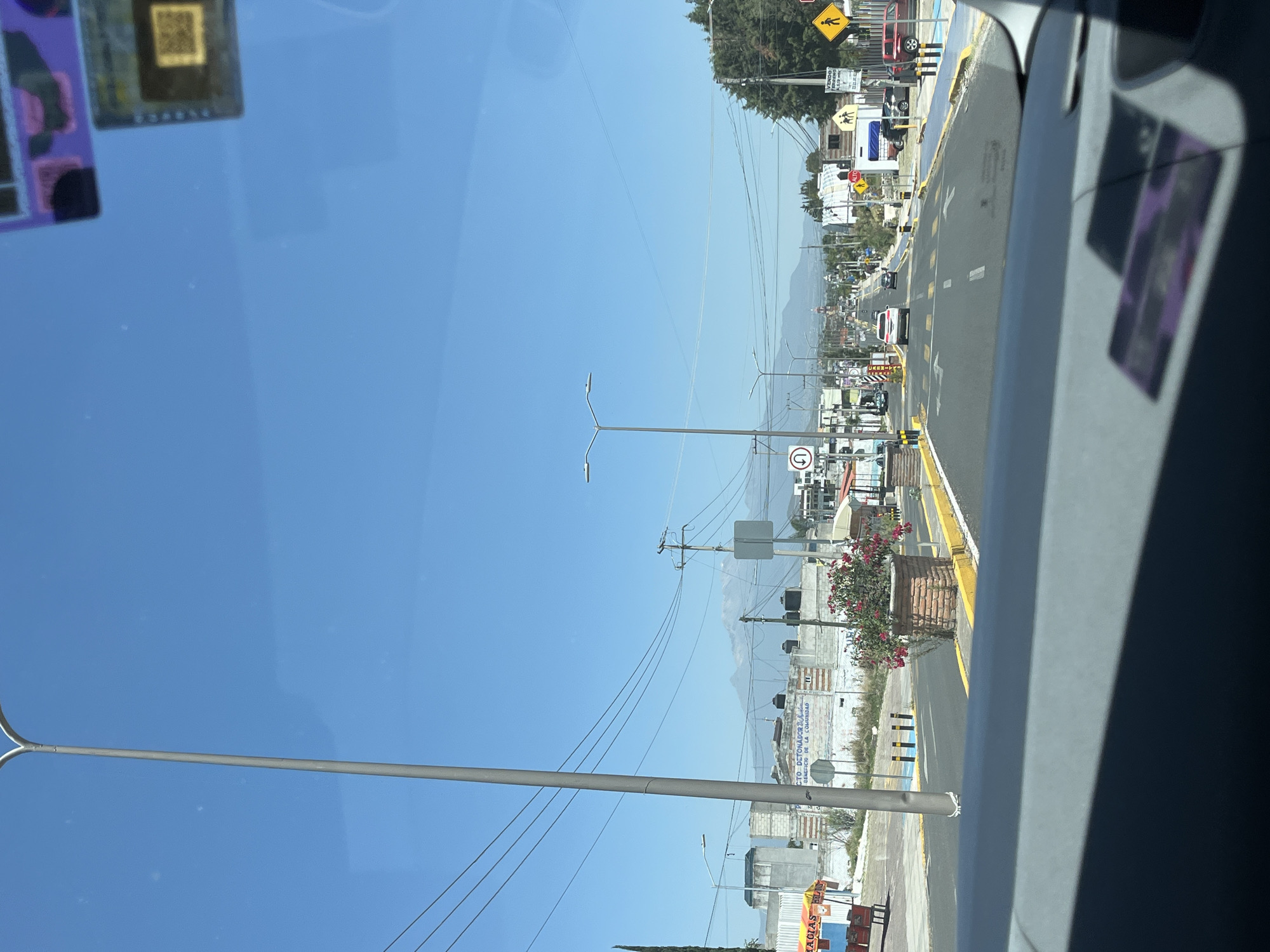 The image captures a bright, sunny daytime scene from the perspective of inside a vehicle, looking down a road in San Andrés Cholula, Mexico. The sky is clear and blue, dominating the upper left portion of the frame.
In the immediate foreground, on the left, reflections from the vehicle's interior are visible, including several purple stickers and a small gold square with a QR code.
The main subject is a multi-lane road stretching into the distance, marked with clear yellow lines and white arrows indicating traffic flow. Several vehicles, including a red SUV and a white sedan, can be seen at various distances along the road. Streetlights and utility poles with power lines line both sides of the thoroughfare.
Along the right side of the road, various buildings, possibly commercial establishments or residences, are visible. Green trees and bushes with vibrant pink flowers add splashes of color to the roadside landscape. Distant mountains or hills rise under the clear sky, forming the background and outlining the geographical setting.
Several road signs are present, providing navigational and safety information. These include a yellow diamond-shaped sign depicting a pedestrian, a yellow square sign clearly displaying "ALTO" (Stop), and a circular sign indicating a "No U-Turn" (a U-turn arrow crossed out). Other visible text on signs includes "VENDO" (Sell), partial text like "CTO. DETONADOR..." and "BENEFICIO DE LA COMUNIDAD," and another sign reading "GRACIAS."
The overall scene suggests a typical day on a main road in a semi-urban or suburban area of San Andrés Cholula, with traffic moving and local businesses lining the street, all under a bright sun.