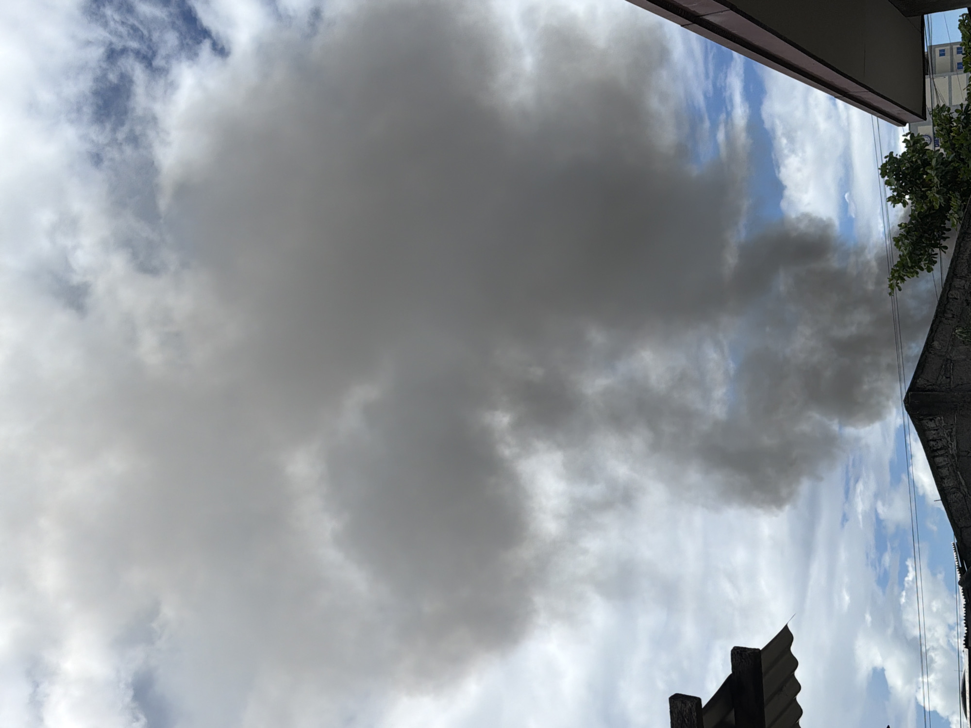 On a cloudy day in Davao City, Philippines, a large plume of dark gray smoke billows into the sky from a source near ground level, partially obscuring the view of the sky. In the foreground, the corrugated metal roof of a building is visible on the left, and the edge of a brown-tiled roof of another building is on the right. Various structures, including what appears to be a concrete wall and the rooftops of other buildings, are visible below the smoke. Electrical wires stretch across the scene, and some green foliage from trees is present. The overall impression is one of an incident involving smoke, possibly from a fire, occurring within an urban or semi-urban environment.