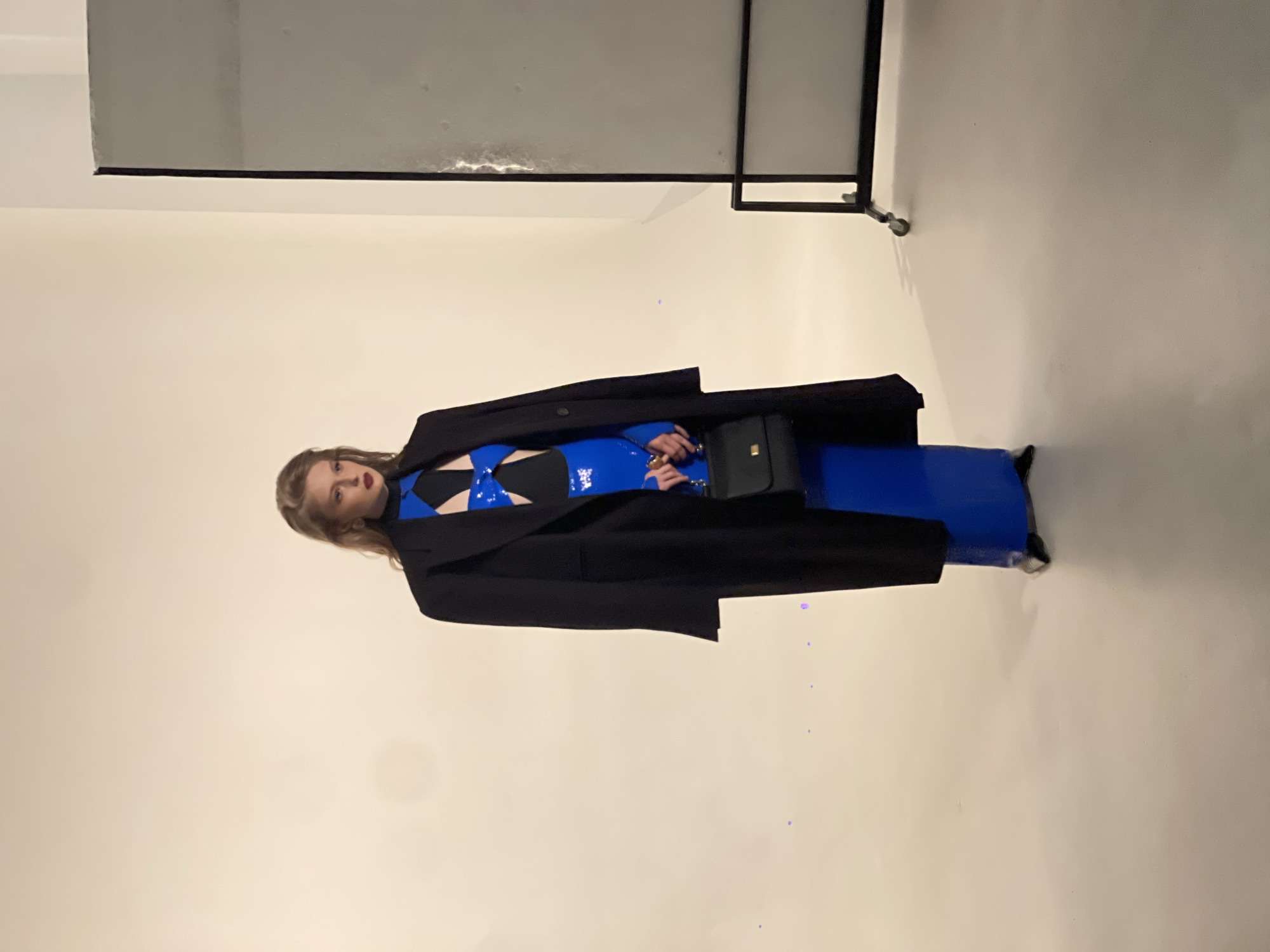 A woman with blonde hair and dark lipstick stands against a plain, light-colored backdrop. She is wearing a long black coat draped over her shoulders, a blue and black patterned top with a cut-out detail, and a long, form-fitting blue skirt. She is holding a small black handbag in her right hand. To the right of the woman is a tall, rectangular screen on wheels. The floor is a neutral color. The setting appears to be a studio. There is no indication of the city of Kyiv, Ukraine, in the image.