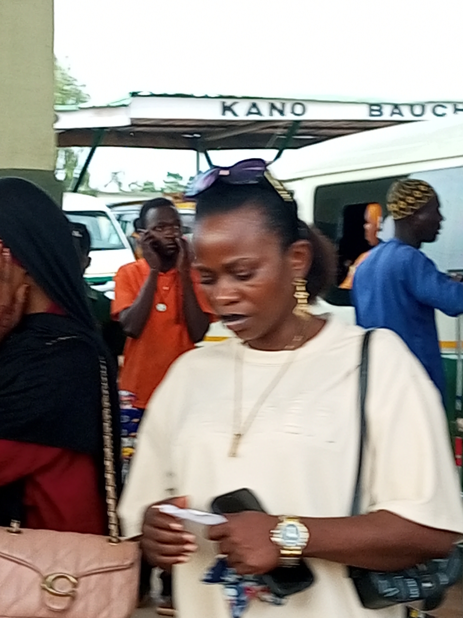 A group of people are gathered outdoors, with a sign overhead that reads "KANO BAUCH". In the foreground, a woman with dark hair pulled back and large hoop earrings is wearing a light-colored t-shirt and holding a white card. She has a watch on her left wrist. To her left, a woman in a black hijab and red shirt is holding a light pink quilted purse. Behind the foreground woman, a man in an orange shirt is on his phone, and to his right, a man in a blue shirt and patterned cap is visible. White vehicles are parked behind the people. The overall setting appears to be a public space, possibly a transportation hub or market area.
